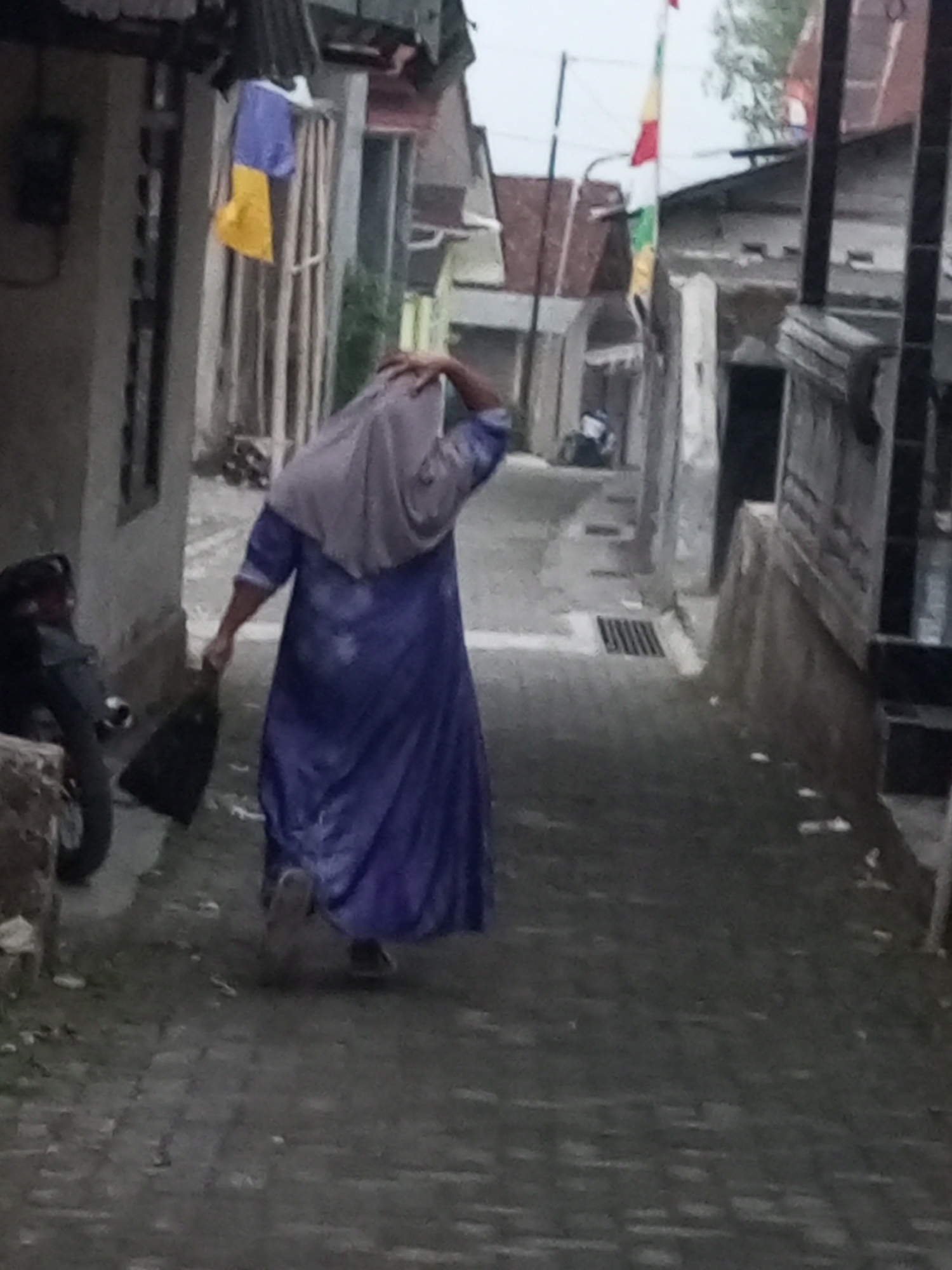 A person walks down a narrow, wet, paved alleyway in Andongsili, Indonesia. The person is wearing a long, flowing purple dress and a light gray hijab that obscures their face. They are carrying a black bag in their right hand. To the left of the alley is a motorcycle. The buildings on either side of the alley are simple structures, some appearing to be made of concrete. A flag with red, white, and yellow sections is visible in the background. The overall impression is that of a utilitarian, residential area in what appears to be overcast weather, given the wet pavement and diffused light.