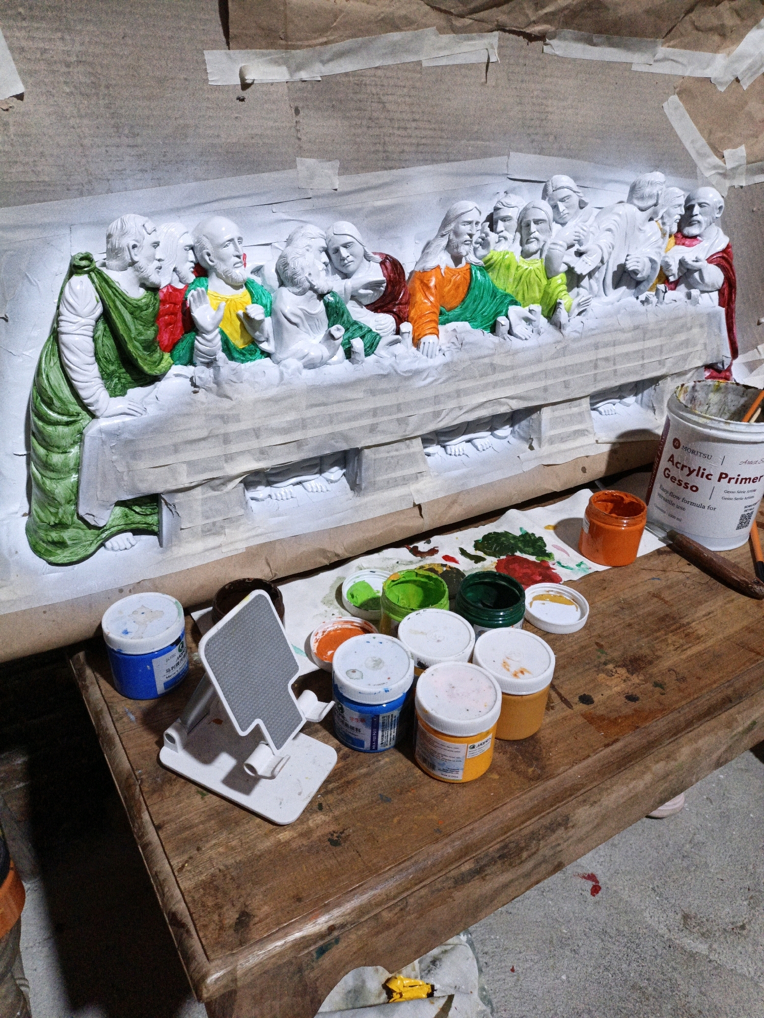 The image shows a plaster cast of "The Last Supper" being painted in a workshop setting. The figures of Jesus and the apostles are partially colored with vibrant hues of green, yellow, orange, and red, while the rest of the cast remains white. Several containers of acrylic paint, primarily in small tubs and a larger can labeled "Moritsu Acrylic Primer Gesso," are arranged on a wooden table in the foreground. A white phone stand is also present on the table. The background depicts a wall covered with brown paper and masking tape, suggesting an ongoing art project. The overall environment appears to be a workshop or studio. There is no indication of a specific location in Cikal, Indonesia, within the image itself.