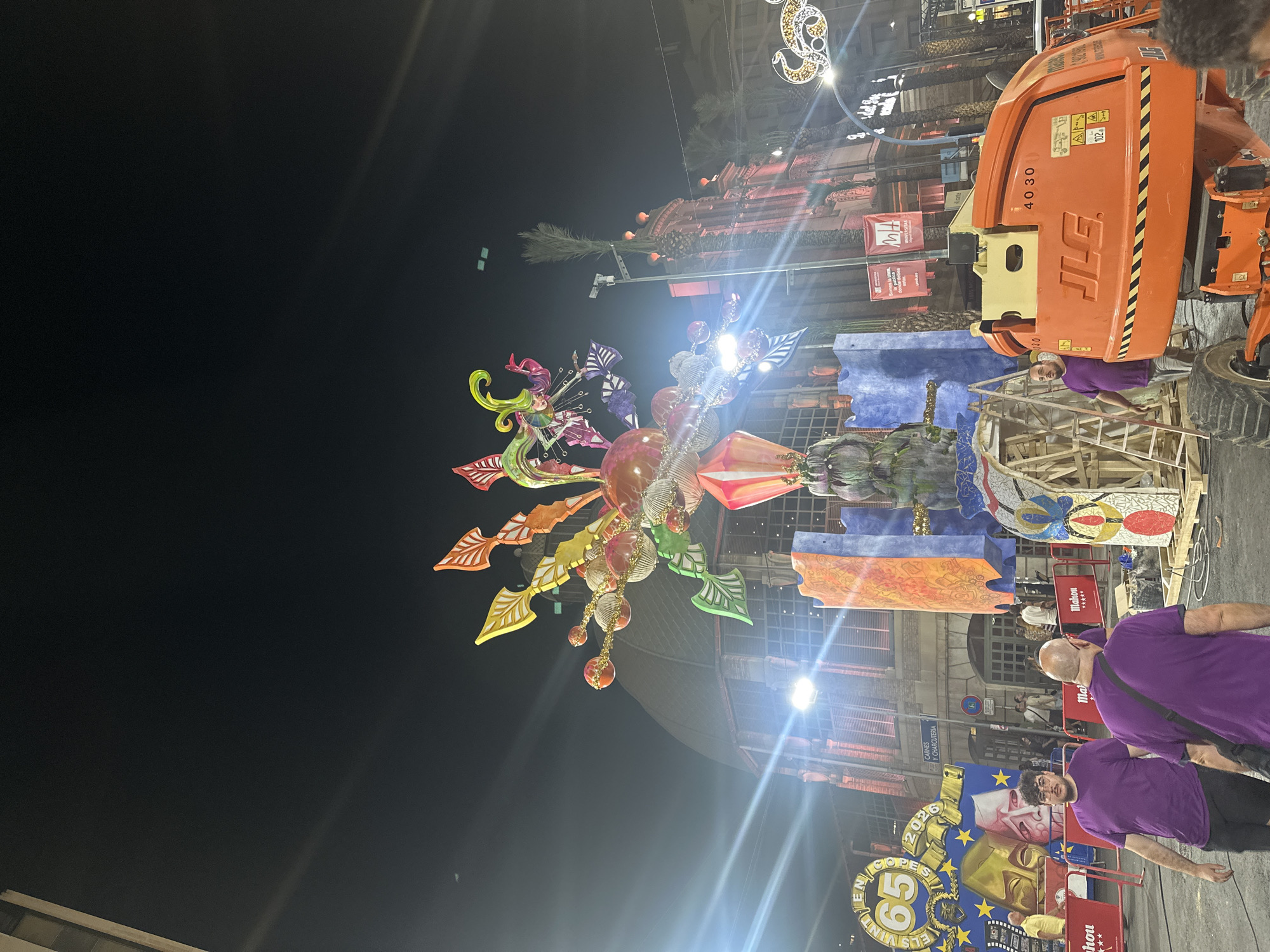 This is a night-time, eye-level, medium shot of an outdoor event, likely a festival, in Alicante, Spain. The dominant subject is a large, colorful, and elaborate illuminated sculpture or float, featuring abstract, organic shapes, spheres, and arrow-like elements in bright reds, oranges, yellows, greens, and purples. This structure is positioned in the foreground and mid-ground, with its base constructed of wooden scaffolding and colorful mosaic-like panels.
To the left of the sculpture, a large banner displays "65" in a golden circle, surrounded by stars and masks, and text in Spanish that includes "2023". A building with a dome and arched windows is visible behind the sculpture, illuminated with red and yellow lights. Several people are present in the scene. In the foreground, two men in matching purple t-shirts and dark shorts are walking, with one facing the camera and the other turned away. Further back, other individuals are seen walking around the event area, some near more scaffolding and banners.
To the right, a large, bright orange aerial work platform (JLG 4030) is parked, with its mechanical arm extended. Behind this, a building with ornate architecture and red lighting is visible, along with palm trees and streetlights. Christmas-like lights in a swirling pattern are also visible in the upper right corner. The overall atmosphere suggests a festive and public gathering at night. The presence of the "65" and "2023" suggests a significant anniversary event. The architecture of the dome-like building and the style of the street lights are consistent with Alicante.
