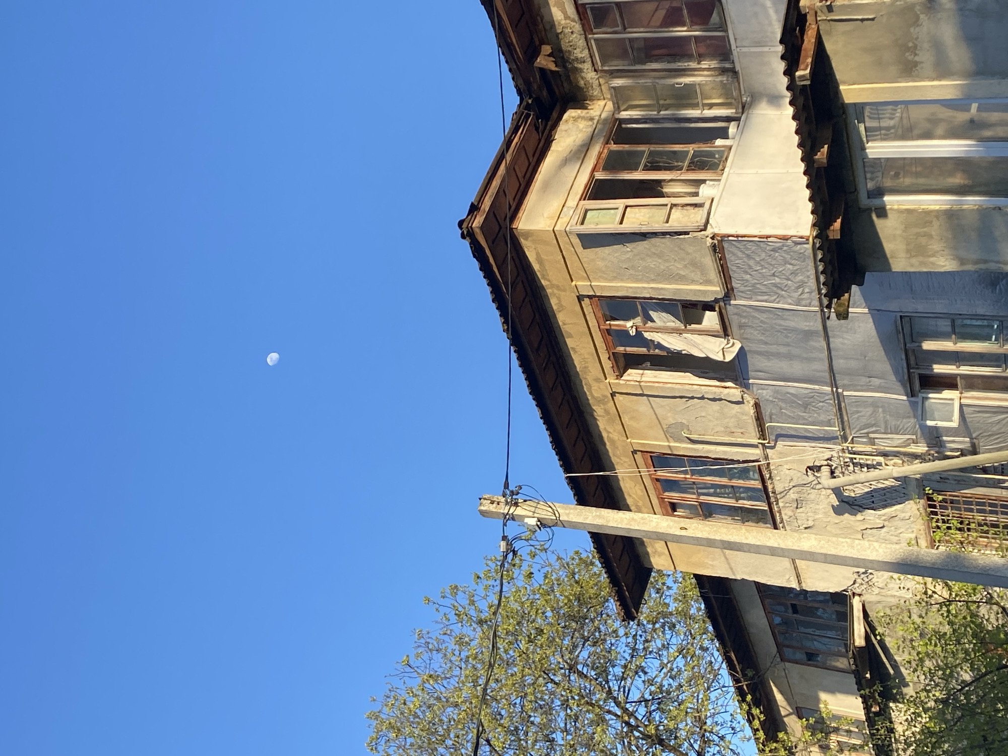 The image shows a three-story residential building under a blue sky with a visible moon. The building appears aged and in disrepair, with weathered concrete and deteriorating window frames. Some windows are covered with makeshift materials. An electrical pole extends from the building, with wires and a tree obscuring part of the structure. The building seems to be in Sochi, Russia.
