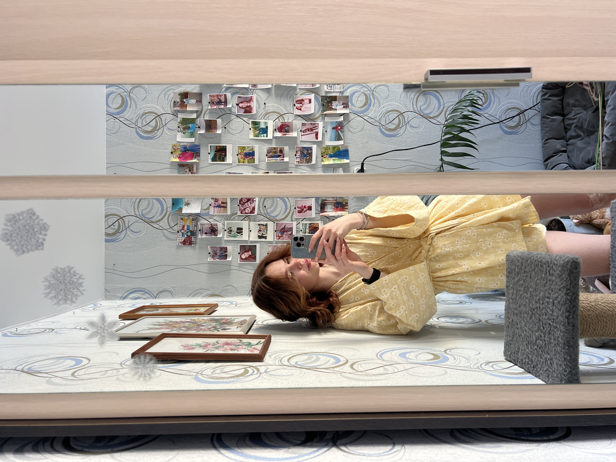 A young female, appearing to be in an indoor residential setting within Chaikovsky, Russia, is captured in a selfie reflected in a mirror. She possesses shoulder-length, wavy brown hair with lighter highlights, and is wearing a yellow floral-patterned wrap-around robe or dress. Her left wrist displays a black band, possibly a smartwatch, and her right hand holds a light blue smartphone, which she is using to capture the image. Her gaze is directed towards the phone screen.
The room features light-colored wallpaper adorned with abstract blue and brown swirling patterns. On the wall behind her, several framed pictures hang, and decorative white snowflake ornaments are affixed to the wall and ceiling. To her right, a collection of small, informal photographs, potentially instant prints, are displayed on the wall, arranged with clips and strings. A black electrical cable is visible trailing down the wall among these photos.
In the foreground, a multi-tiered cat scratching post or tree is visible, featuring a grey carpeted platform and sisal rope sections. Reflected in the mirror to the right of the woman, a dark grey pet bed or small sofa is partially visible, along with the leaves of a green houseplant. The mirror itself is part of a light-colored wooden wardrobe or door frame. The scene suggests a personal living space.