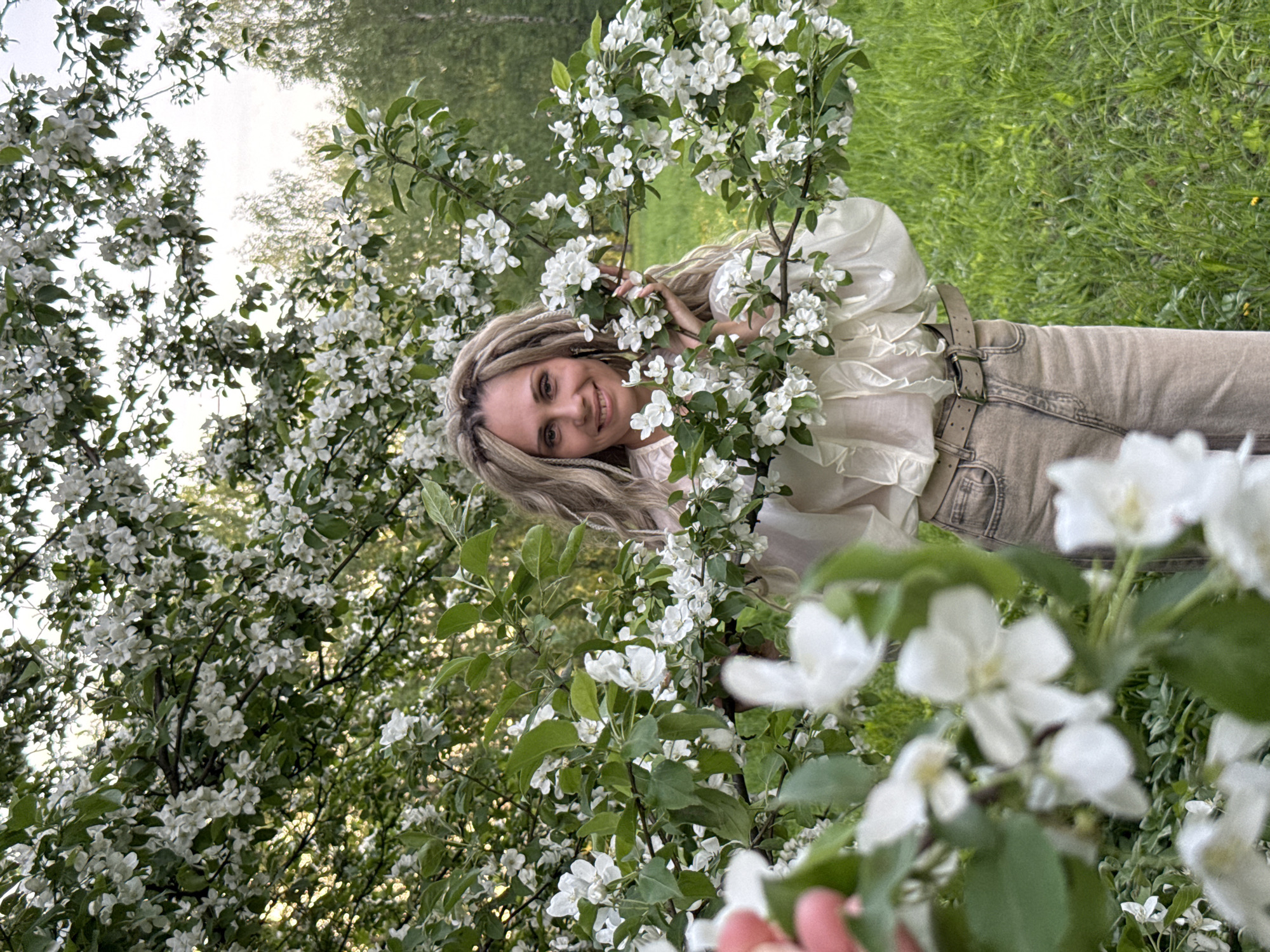 A woman with blonde dreadlocks stands in the town of Шебунино, Russia among apple blossoms in a sunny outdoor environment. She is wearing a cream-colored blouse with ruffles and light brown pants. She smiles, her hands delicately positioned among the white flowers and green leaves. Lush green grass forms the backdrop behind the blossoming tree.