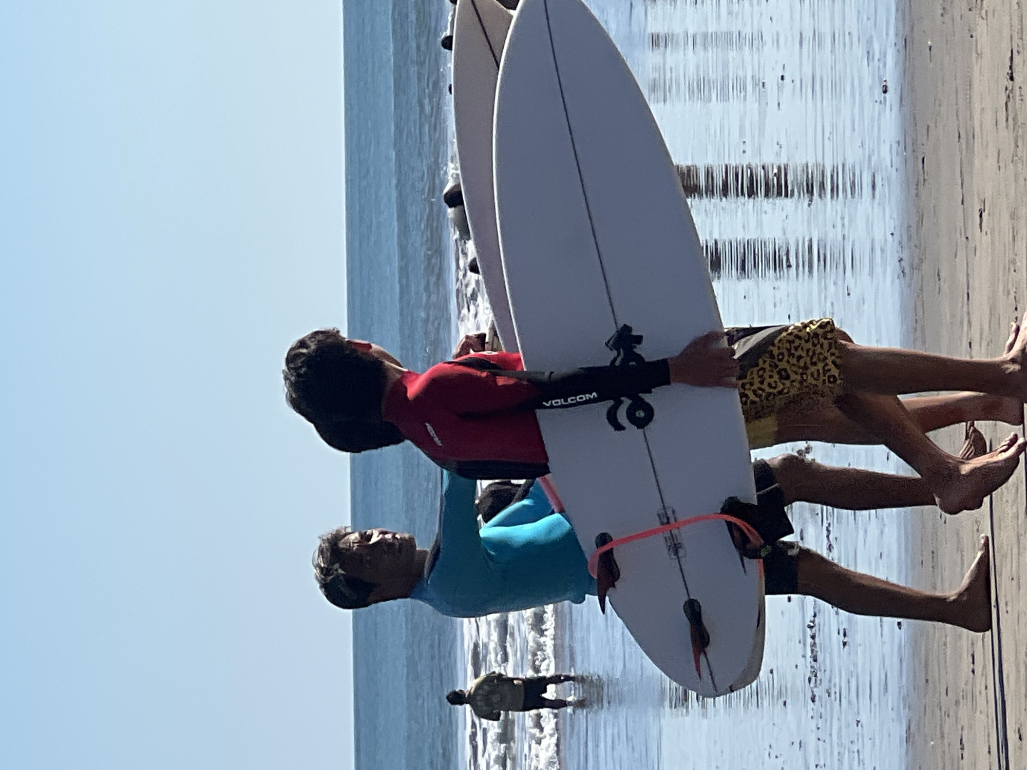 Two young men are walking on the beach in Kuta, Indonesia, carrying surfboards. The man in the foreground is wearing a bright blue long-sleeved rashguard and black shorts. He is looking to his left, towards the ocean, with his right arm extended. He is carrying a white surfboard with a pink leash. Next to him, a second young man is wearing a red and black long-sleeved rashguard and leopard-print shorts. He is facing to his right, and his left arm is obscured by the first man's surfboard. He is also carrying a surfboard, partially visible behind the first. In the background, a third man is standing in the shallow water, facing away from the camera, wearing a light-colored shirt and dark shorts. The ocean is visible, with gentle waves breaking on the shore. The sand is wet from the receding tide, and reflections of the sky and the surfers are visible on the water's surface. The sky is clear and blue.