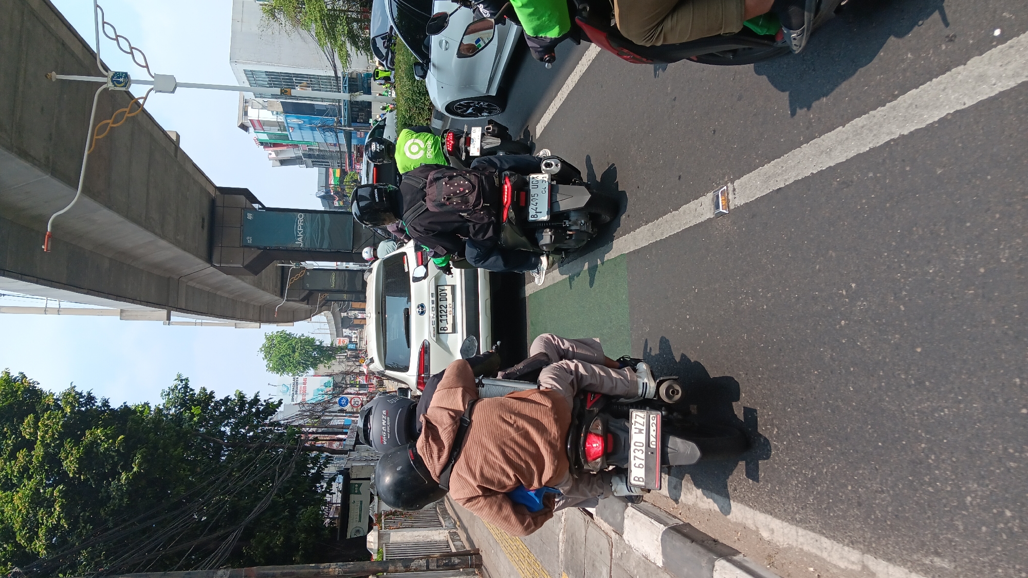 This is a full, eye-level, vertical shot of a busy street scene in South Jakarta, Indonesia, likely during daylight hours as indicated by the bright sunlight casting shadows. The foreground features two individuals riding a scooter, with the passenger in the rear wearing a brown hoodie and grey pants. The driver of this scooter is wearing a black helmet. They are in the middle of a lane, on asphalt road, bordered by a sidewalk and a raised green lane marker. Behind them, a white SUV with license plate B 1122 DOY is in traffic, followed by another scooter with a rider wearing a green jacket and black helmet. Further down the road, more vehicles and buildings line the street, indicating an urban environment. An elevated structure, possibly an overpass or a highway ramp, dominates the upper portion of the image, with its concrete supports and signage. The overall atmosphere suggests a typical day of traffic in a densely populated area.