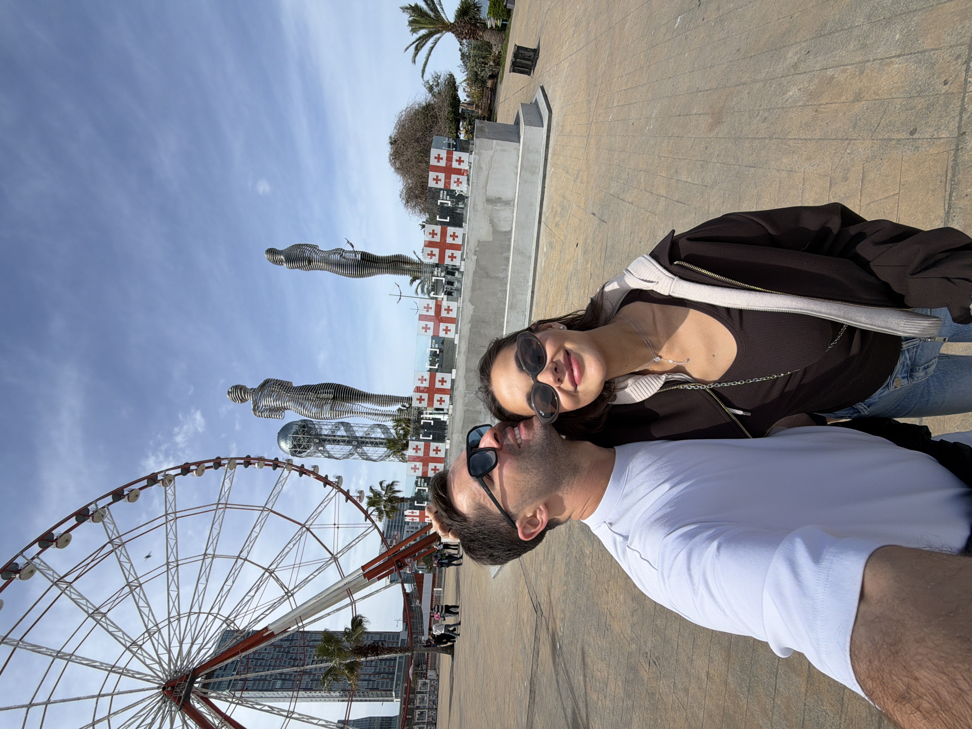 A selfie captured in Batumi, Georgia, shows a couple smiling. The woman has long brown hair, sunglasses, a dark brown top, and a light-colored hoodie. The man, wearing a white t-shirt and sunglasses, has short dark hair and a beard. In the background is the Batumi Ferris Wheel, the "Ali and Nino" statue, and Georgian flags. The ground is tiled. The sky is partly cloudy.