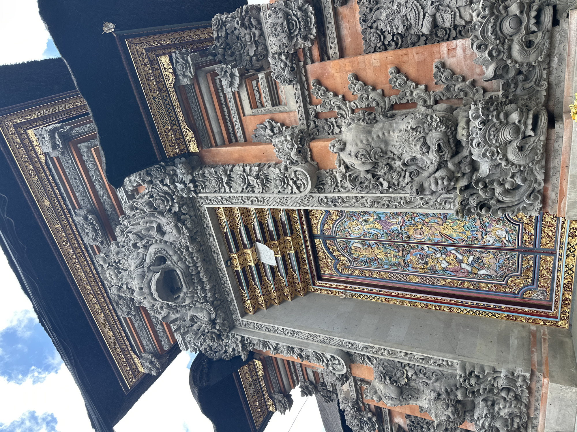 The image depicts the intricately carved facade of a temple in Ubud District, Indonesia. The temple features a large, ornate doorway with colorful, detailed paintings. Flanking the doorway are stone carvings of mythical creatures and floral motifs, with prominent gargoyle-like figures above the entrance. The structure has a traditional thatched roof. The sky above is partly cloudy, suggesting daytime. The overall impression is of a highly decorative and culturally significant architectural site.