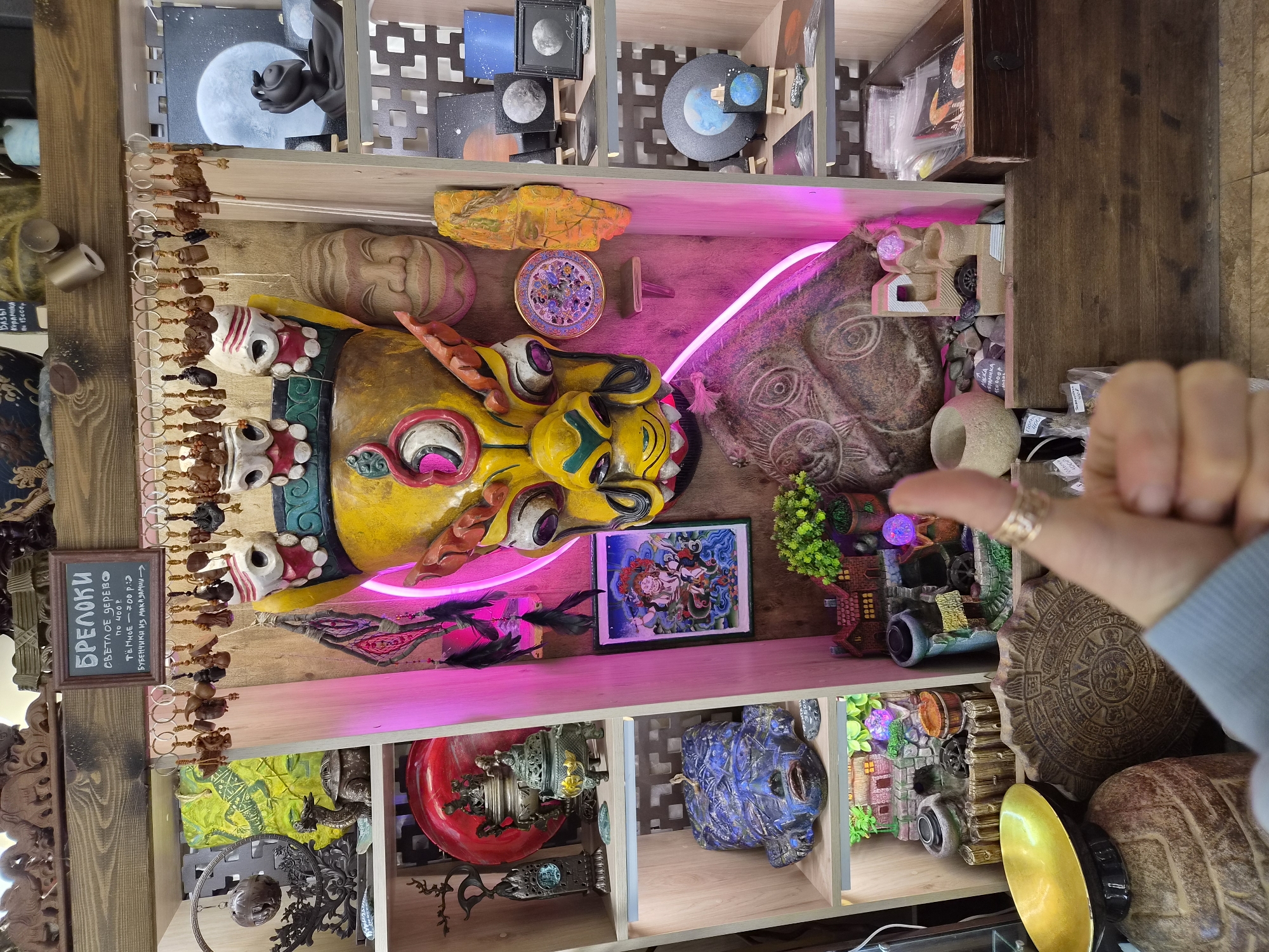 The image displays a close-up view of a shelf filled with various decorative and artistic items, likely in a shop or market. A hand with a ring is giving a thumbs-up gesture in the foreground, partially obscuring some of the displayed items.
The shelf is divided into several sections. On the left side, there are decorative plates, incense burners, and a blue, sculpted mask with facial features. In the center, a large, brightly colored, multi-faced mask with yellow and red accents is prominent. Below this mask are smaller decorative houses and plants, as well as a stone carving of a circular calendar-like object and a golden bowl. To the right of the central mask, there are carved wooden faces, and further to the right, a series of shelves display small paintings and circular art pieces depicting the moon and planets, as well as what appear to be small ceramic items. A wooden drawer filled with packaged items is visible at the bottom right.
Above the central mask, a sign with Russian text reads "БРЕЛОКИ" (keychains), with further details indicating prices and materials. A collection of various keychains hangs from a bar above the sign.
The overall environment appears to be indoors, with wooden shelving and textured walls. A neon pink light illuminates parts of the central display. The items suggest a theme of cultural artifacts, possibly with an Eastern or mystical influence, given the masks and figurines.
Based on the Russian text on the sign and the context of such displays, this scene is likely located in a souvenir shop or a market selling handicrafts in the city of Vladivostok, Russia.