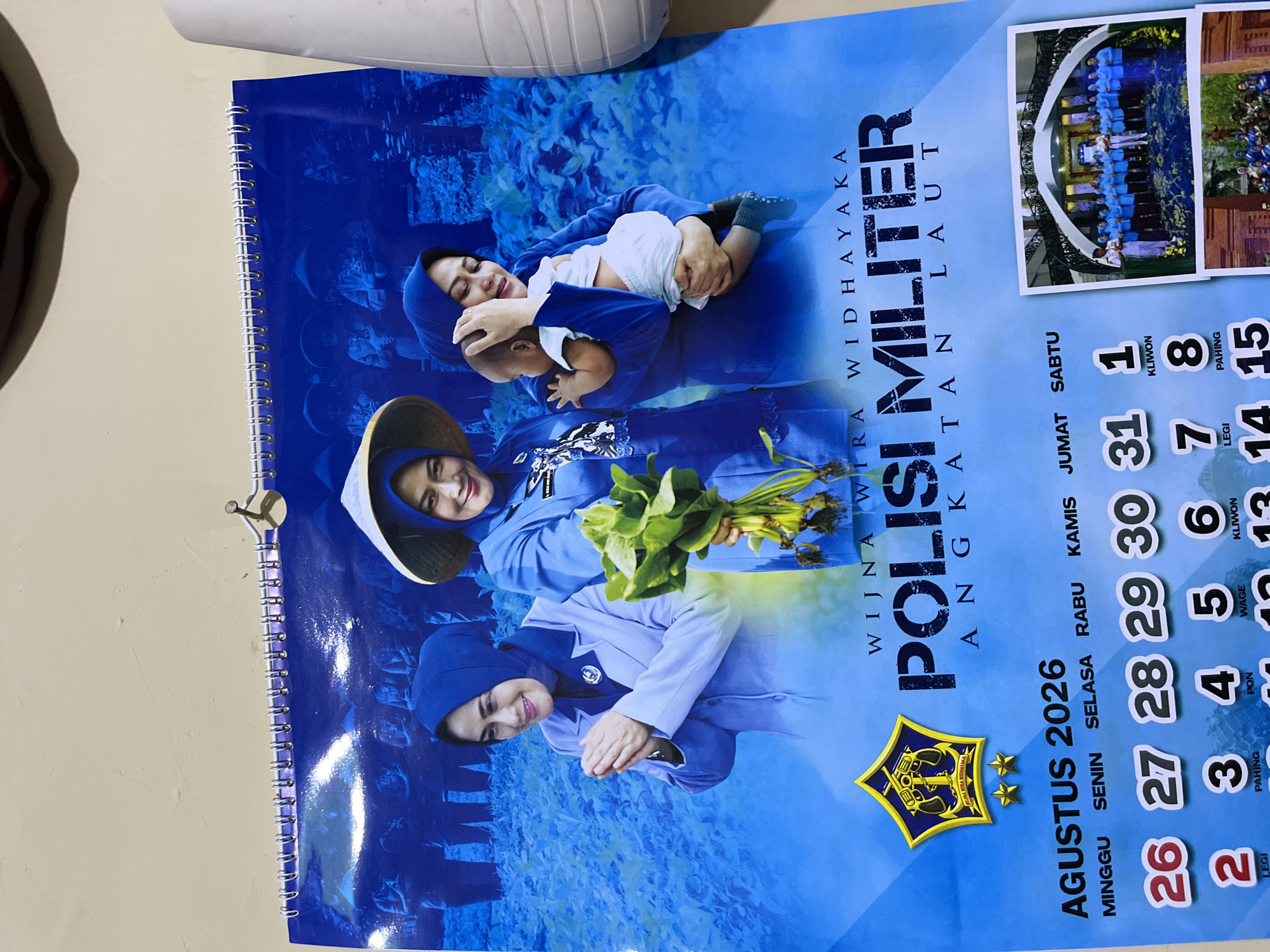 This is a photograph of a wall calendar for August 2026. The calendar features a blue background with images of women in uniform. The central image shows three women, two of whom are in blue uniforms and hijabs. One woman on the left is holding her hands together in a gesture of greeting, and the woman in the center is holding a baby. The third woman, on the right, is also in uniform and holding a bouquet of green leaves. The calendar text reads "AGUSTUS 2026" and lists the days of the week in Indonesian: "MINGGU," "SENIN," "SELASA," "RABU," "KAMIS," "JUMAT," and "SABTU." Below the days of the week, there is a grid of numbers representing the dates, with some dates having additional text in Indonesian such as "LEGI," "PAHING," "PON," and "KLIWON." The title of the calendar appears to be "POLISI MILITER ANGKATAN LAUT." The calendar is hanging on a wall, attached with a spiral binding. There is a small nail visible at the top of the calendar. To the right of the calendar, a white object, possibly a vent or part of a wall fixture, is visible. The lower portion of the photograph contains two smaller inset images, one showing a group of people in uniform standing in a hall, and another showing a group of people in uniform outdoors. The location is identified as Dengkol, Indonesia.