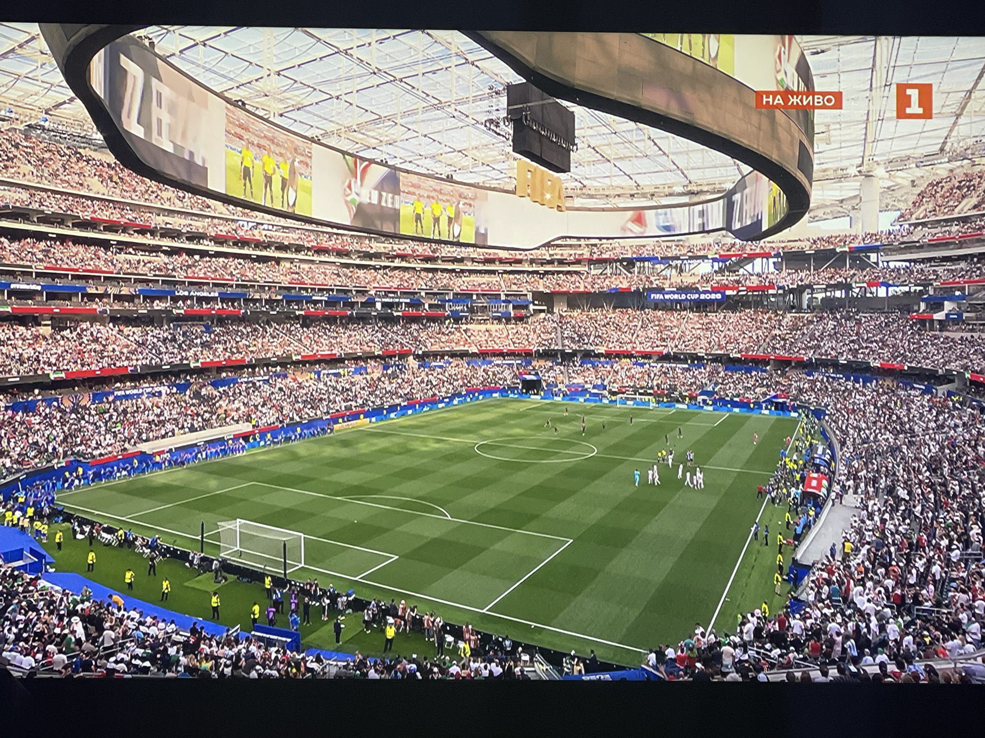 This image depicts a wide-angle view of a crowded soccer stadium during a match. The stadium features a large, curved video screen displaying text and images, including the word "FIFA" in large golden letters and the phrase "НА ЖИВО" (meaning "Live" in Bulgarian), along with a graphic of the number "1". The soccer field is brightly lit and striped with green and white lines. Numerous spectators are seated in the tiered stands surrounding the field, many wearing white and red. Players in white and dark uniforms are visible on the field. Security personnel in yellow vests are positioned around the perimeter of the field and among the crowd. The stadium appears to be a modern, open-air venue with a visible roof structure. There is no indication of the city of Ruse, Bulgaria, in the image or its context.