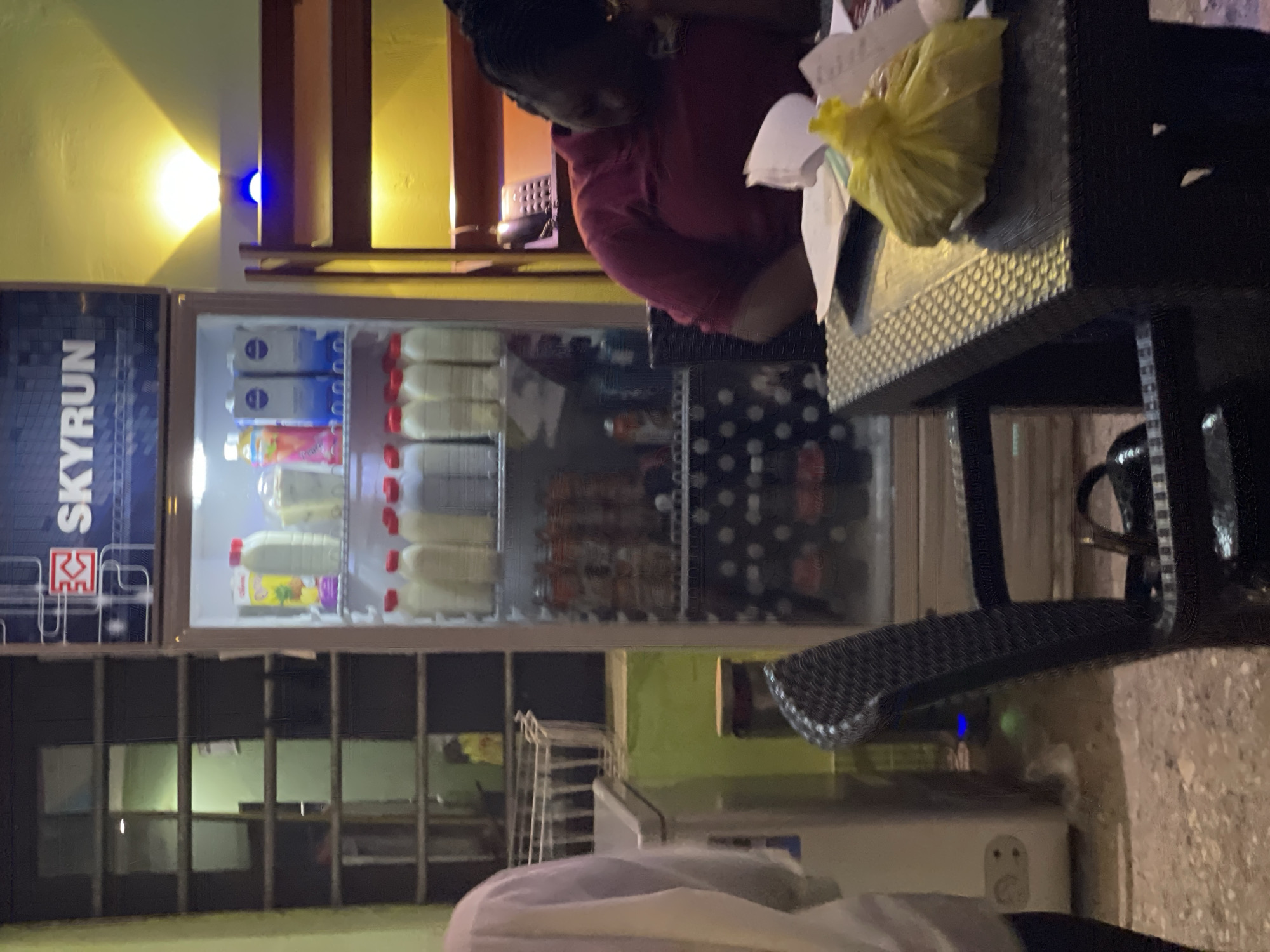 Akwanga, Nigeria. Indoor view of a store or business. A woman is seated at a table laden with papers and a yellow plastic bag. Behind her is an open refrigerator stocked with bottled and packaged beverages. A "Skyrun" sign is visible on the side of the refrigerator. Behind the refrigerator is a set of shelves and a freezer. The environment is dimly lit.