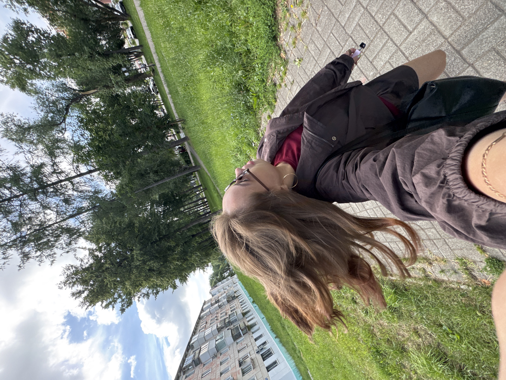 In Chaikovsky, Russia, a young woman with long, light brown hair styled to fly outward is captured in a selfie on a paved sidewalk. She is wearing sunglasses, a red shirt, and a dark brown jacket, with a black bag slung over her shoulder. The background features a multi-story apartment building with light-colored walls and numerous balconies on the left, and a grassy area with a line of trees on the right. The sky is cloudy with patches of blue. The perspective is low-angle, suggesting the photo was taken from a low vantage point.