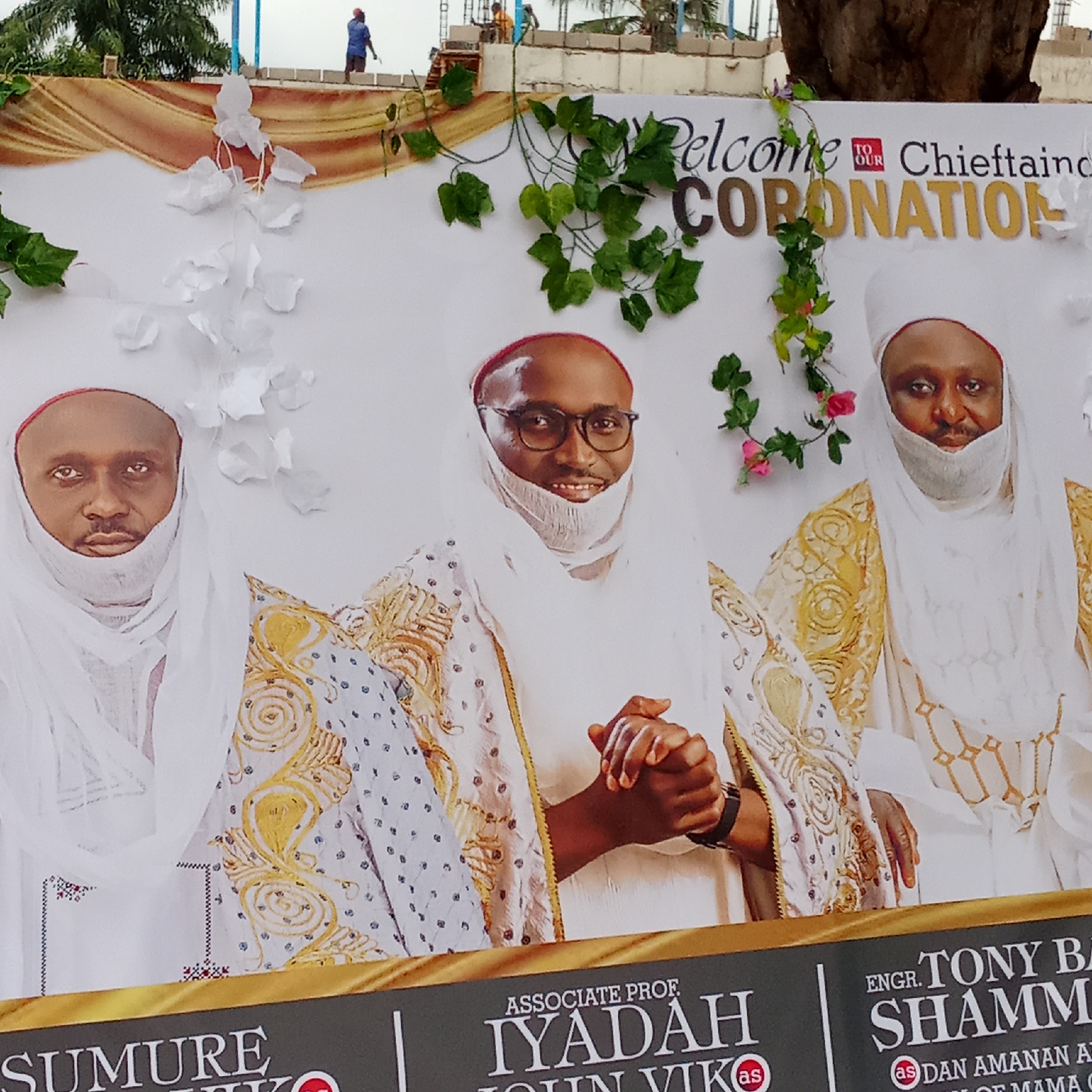 The image shows a banner that reads, "Welcome to Our Chieftaincy Coronation." Below this text are portraits of three men in traditional garb. The middle man wears glasses and has his hands clasped in front of him. Below the portraits are printed names and titles. From left to right, they read "SUMURE VIK as," "ASSOCIATE PROF. IYADAH JOHN VIK as," and "ENGR. TONY BA SHAMM as DAN AMANAN A OF MAMA G." The banner is decorated with white flowers and green vines. In the background, above the banner, there is the suggestion of a building under construction and figures working on it. Given the details of the names and titles on the banner, this is likely in Akwanga, Nigeria.