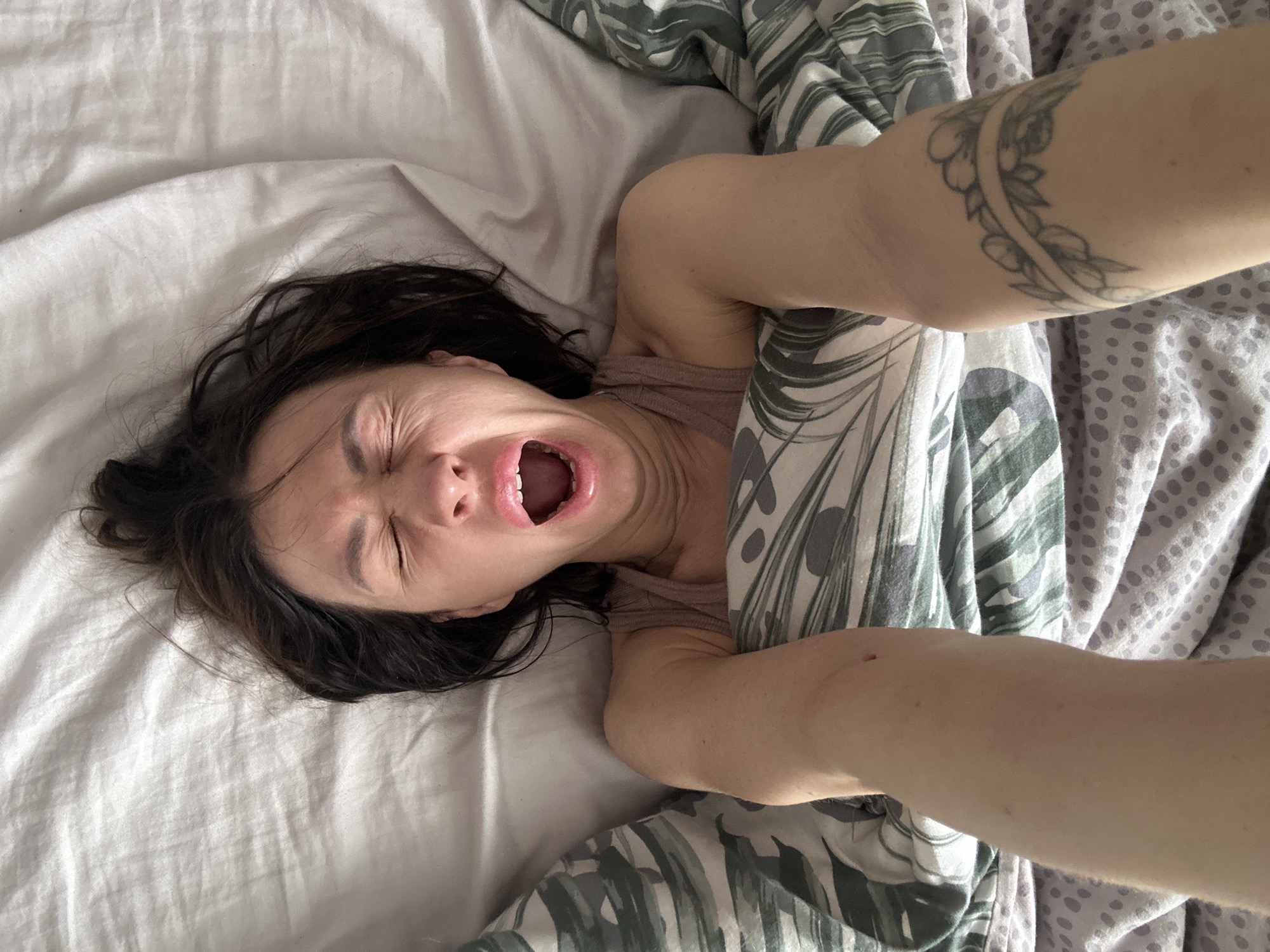 A woman is lying in a bed. She has dark hair and is wearing a light-colored top. Her mouth is open in a yawn or scream. She is surrounded by white and patterned bedding. Her left arm is visible and has a tattoo. The location is unknown.