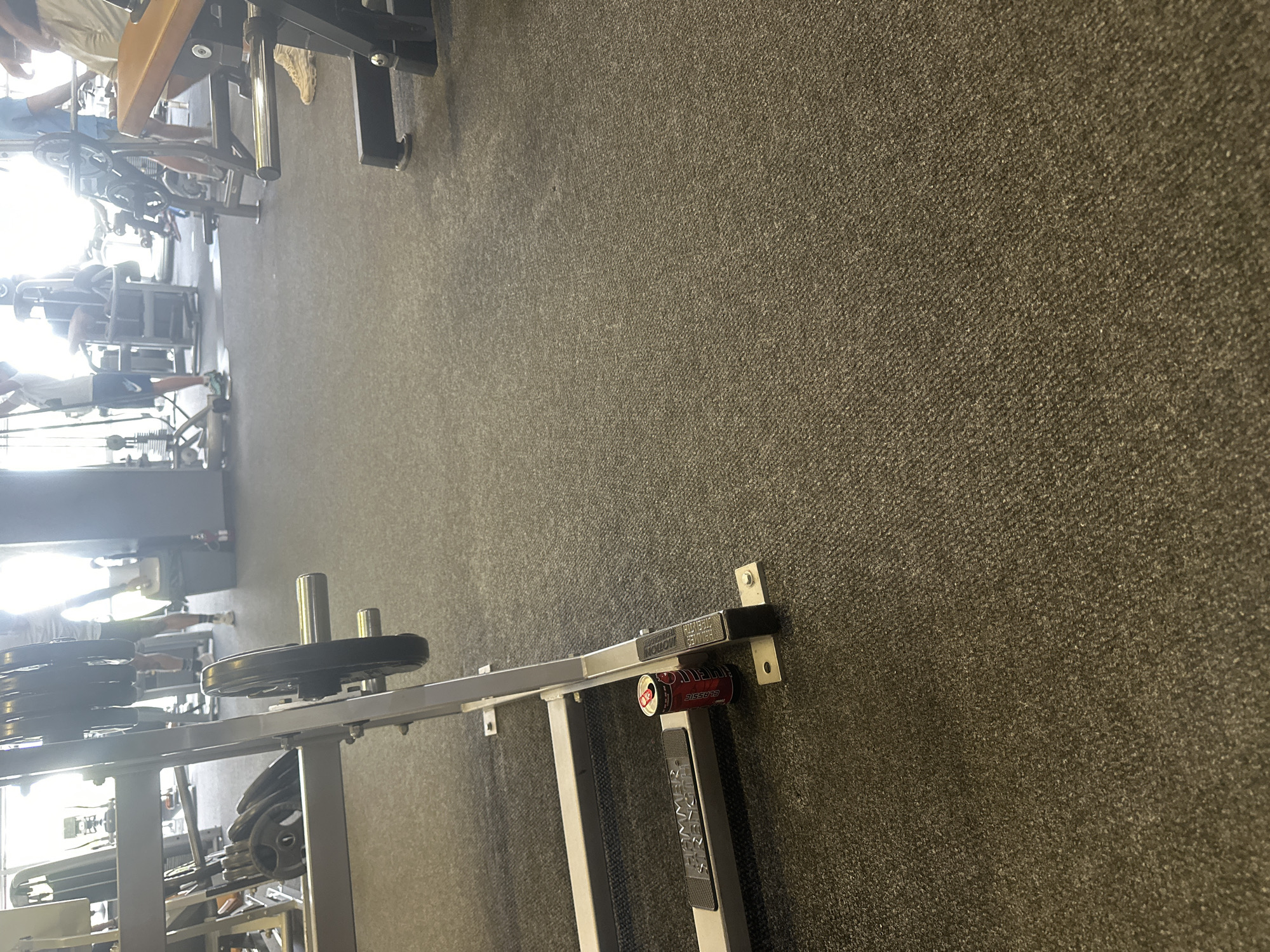 The image, taken in Blagoevgrad, Bulgaria, presents a dynamic scene within a brightly lit gym, rotated 90 degrees clockwise. The perspective is slightly elevated, looking across a carpeted floor towards various pieces of exercise equipment and people.
In the mid-ground and background, several individuals are actively engaged in their workouts. They are seen using different gym machines, including what appear to be leg press or similar strength training apparatuses, indicating a focus on lower body exercise. The individuals are dressed in casual athletic wear, such as shorts, t-shirts, and sneakers. Bright natural light streams into the facility, likely from windows, suggesting it is daytime.
The foreground showcases a sturdy metal gym equipment rack, holding several black and metallic weight plates. A distinct detail is a red beverage can, possibly an energy drink or soda, wedged into a structural component of this equipment. Visible text on the can reads "CLASSIC," while the gym equipment itself displays the brand name "HAMMER STRENGTH." The entire floor area is covered with a textured, dark grey-brown carpet, providing a uniform surface throughout the training space. The scene conveys an active, functional gym environment during operational hours.