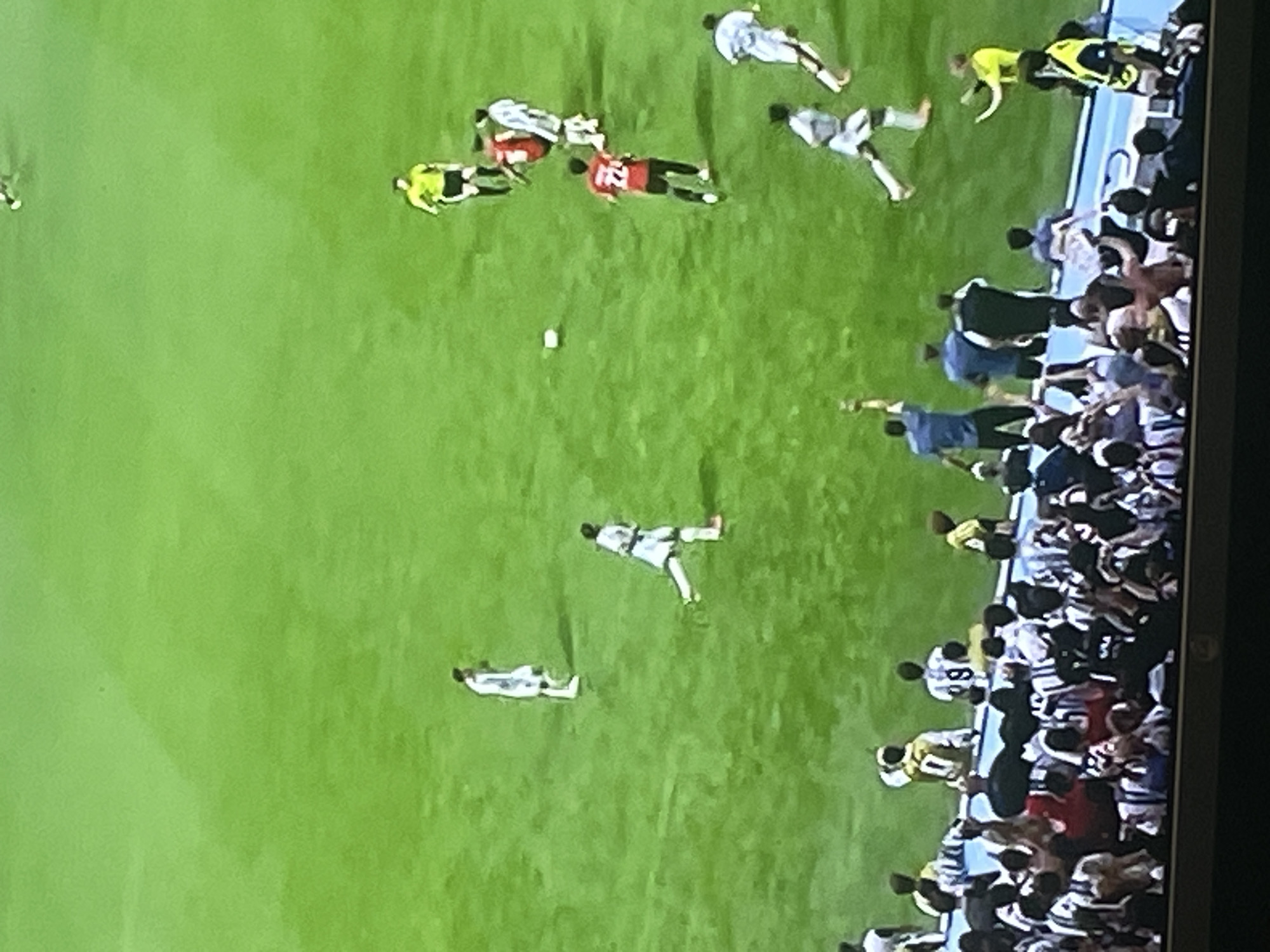 The image captures a soccer match on a bright green field. Several players in white and red uniforms are visible, some in motion, engaged in the game. A referee in a yellow shirt and black shorts is also present on the field. In the foreground, a crowd of spectators is seated in stands, with some individuals standing and appearing to interact with the players or officials. The scene is set during daylight hours, with the field illuminated by natural light. There is no indication of the city of Zhukovo, Russia, in the provided image.