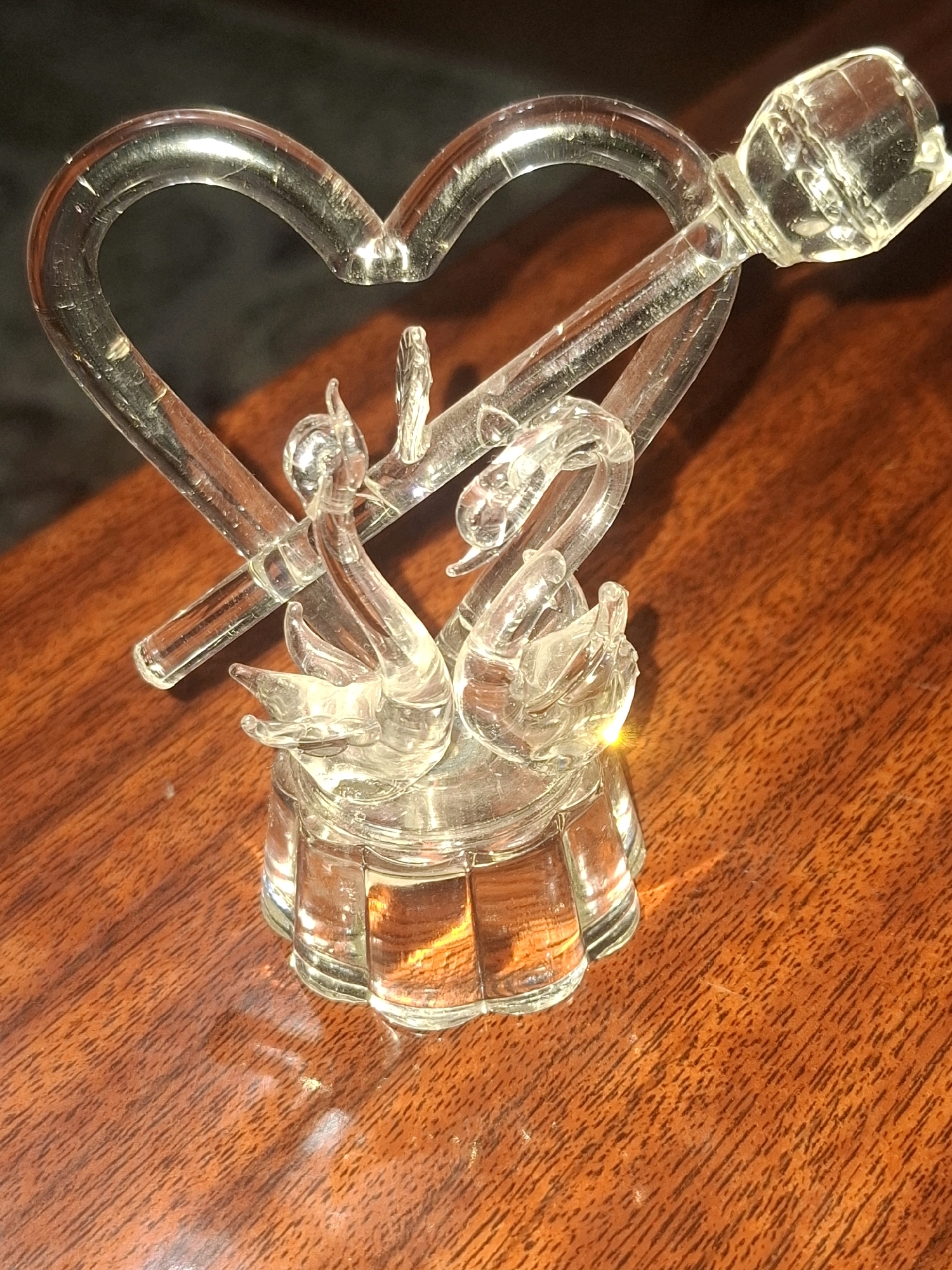 A glass figurine of two swans forming a heart shape sits on a wooden table. The figurine is clear glass and appears to be a decorative item. The table is polished wood with visible grain. The background is dark and out of focus. There are no people or animals present in the image. This image was taken in Almaty, Kazakhstan.
