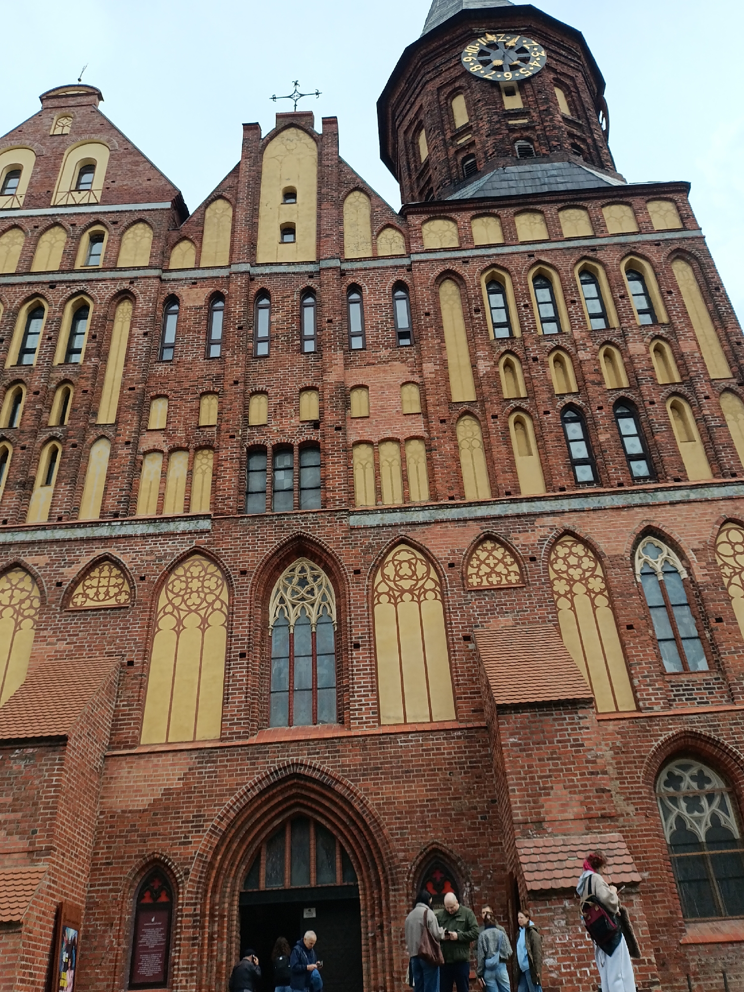 The image depicts a low-angle view of the Königsberg Cathedral in Kaliningrad, Russia. The cathedral is a large brick building with a tower featuring a clock face. The facade has numerous windows, some with elaborate gothic tracery. A group of people stands in front of the cathedral's main entrance. They appear to be tourists, some looking at their phones and talking among themselves. One person stands slightly apart, looking at the building. The sky is overcast, casting a diffuse light over the scene. A sign is visible to the left of the entrance.
