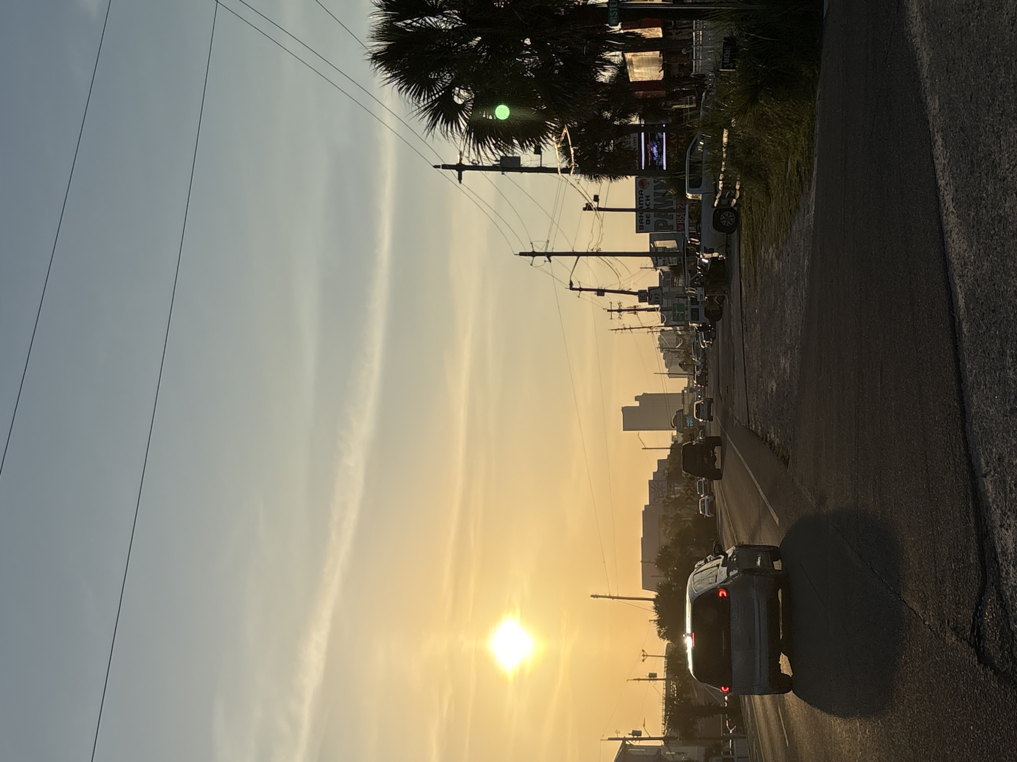 The image depicts a roadside scene, likely a main road or highway, at either sunrise or sunset, indicated by the prominent low sun and long shadows. The overall lighting is a warm, golden hue.
In the immediate foreground on the right, the rear of a silver minivan, possibly a Honda Odyssey, is visible with its brake lights illuminated, suggesting it is either stopped or in slow-moving traffic. Its large shadow extends onto the asphalt road. The road itself appears to be a multi-lane thoroughfare with vehicles stretching into the distance.
Along the left side of the road, numerous utility poles with power lines run parallel to the direction of travel. Several palm trees are scattered along this side, with one large palm tree in the mid-ground obscuring some power lines and buildings. A bright, green circular lens flare or light anomaly is visible within the foliage of this palm tree.
Commercial buildings line both sides of the road. On the left, a sign is clearly visible displaying "BAHAMA BEACH" and another large sign prominently features the word "PAWN," with partial numbers like "4852" also visible. Another sign appears to have "TWIST" on it, but is partially obscured. Taller, multi-story buildings, possibly hotels or condominiums, can be seen in the distant background on the left, silhouetted against the bright sky.
The sky transitions from a light blue at the top to a brilliant golden-yellow closer to the horizon, where the sun is a radiant point of light, creating a strong lens flare. Some faint, wispy clouds are present in the sky. The scene conveys a sense of a busy commercial strip, characteristic of a tourist destination like Panama City Beach.