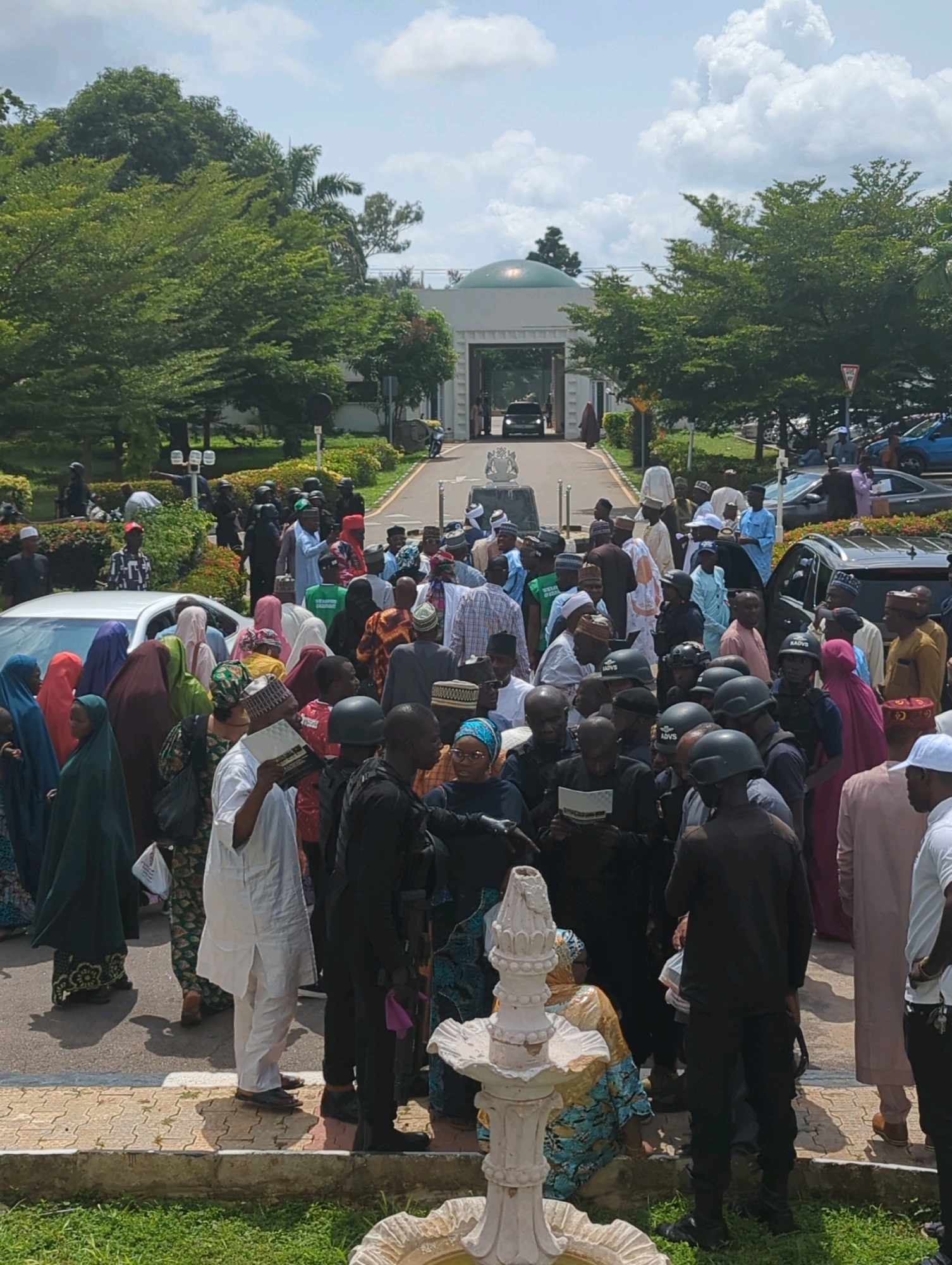 The image depicts a gathering of people, many of whom are wearing traditional Nigerian attire, in front of a white building with a green domed structure visible through an archway. The scene appears to be outdoors, with lush green trees and well-maintained landscaping surrounding the building. A paved walkway or road leads towards the entrance, with vehicles visible in the background. Several individuals are wearing what appear to be security uniforms, some with helmets. The overall impression is of a formal or public event, possibly a reception or ceremony, taking place in a significant location in Unguwan Dosa, Nigeria. The crowd is diverse, with men, women, and possibly children present. Some individuals are holding documents or bags. The lighting suggests it is daytime.