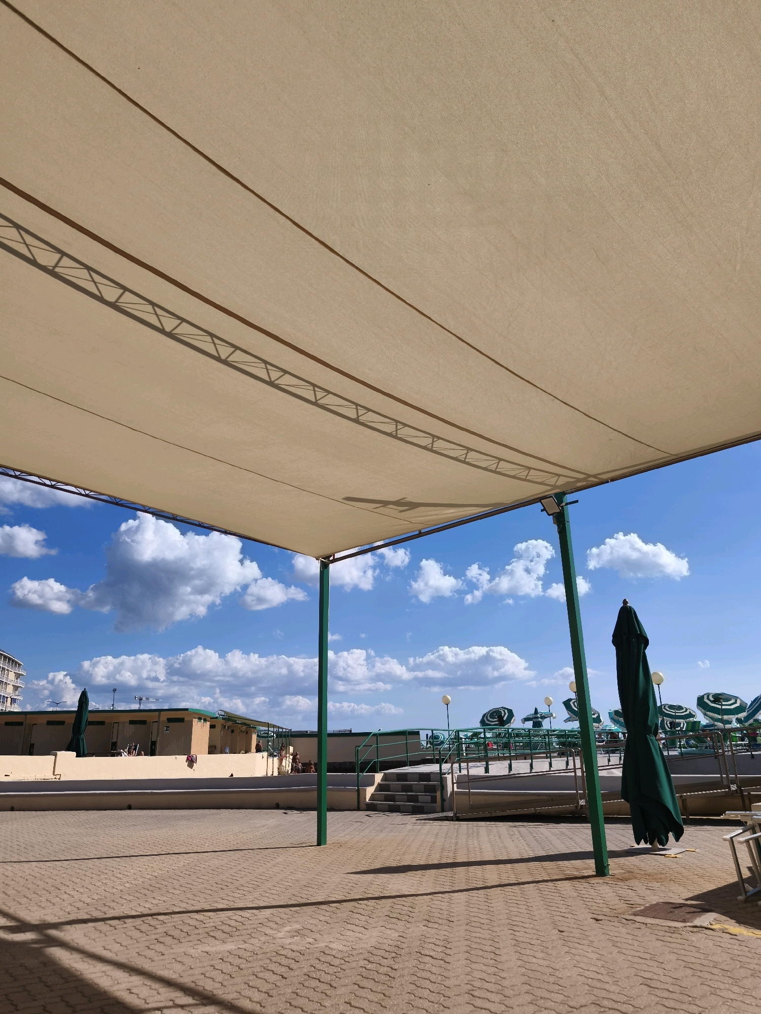 The image captures an outdoor paved area under a large, light-colored canopy supported by green poles. The canopy casts a shadow on the textured pavement. In the background, a bright blue sky is dotted with white, fluffy clouds. Beyond the paved area, there is a row of buildings with tan walls and green trim. Several closed green umbrellas stand upright. Further back, there are more open green and white striped umbrellas, as well as lampposts. The overall impression is of a sunny day at a seaside resort or public beach facility, likely in Livorno, Italy, given the architectural style and the presence of beach amenities. There are a few people visible in the background, some appearing to be seated or walking near the buildings and umbrellas.