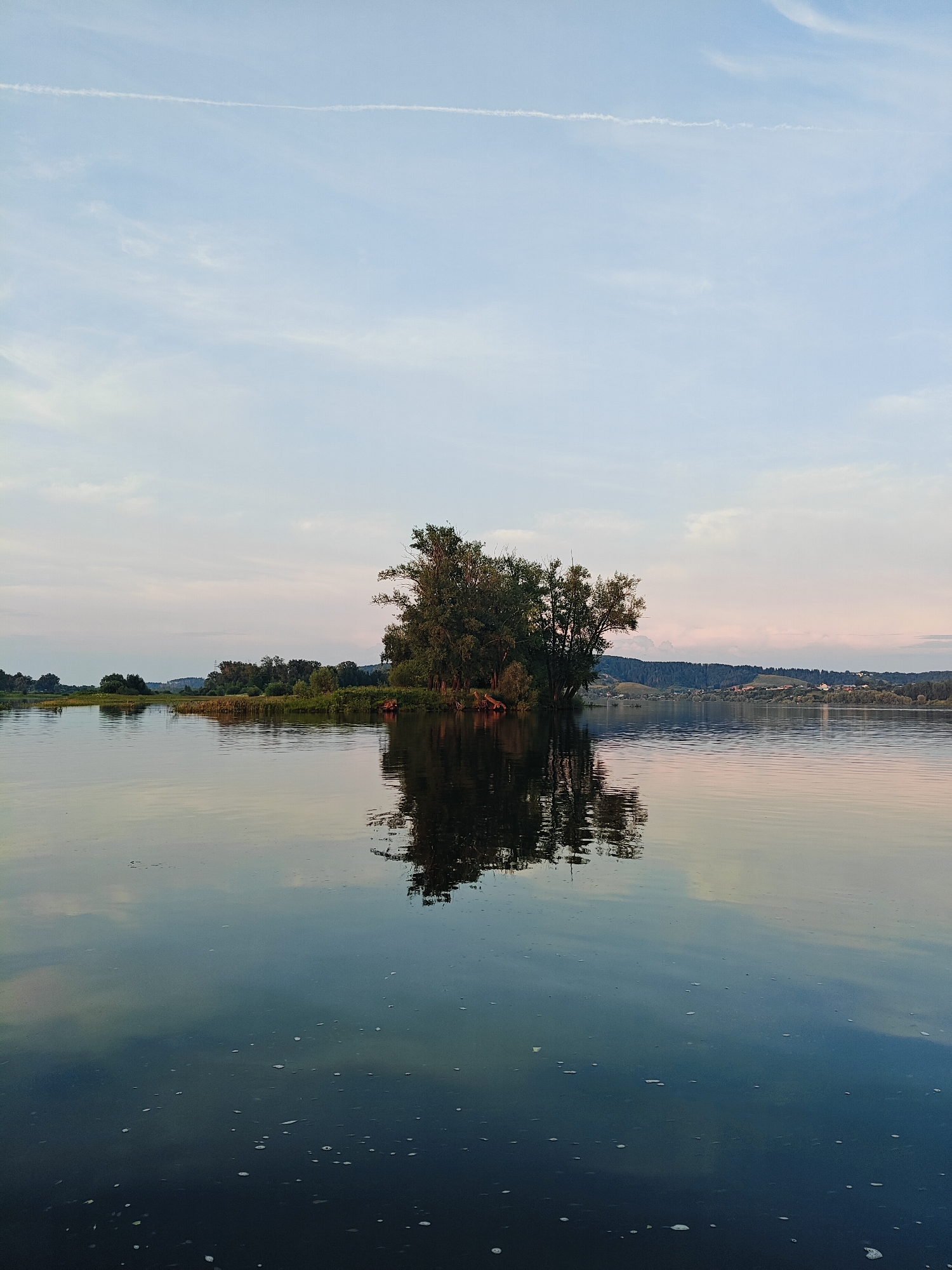 A tranquil scene unfolds on a wide, calm river in Ol'khovskoye, Russia. The water's surface acts as a mirror, reflecting the soft hues of the sky and the lush greenery of the landscape. Dominating the center of the frame is a small, densely wooded island, its reflection a dark silhouette beneath the trees. Along the distant banks, rolling hills covered in trees and dotted with small clusters of buildings suggest the presence of a rural settlement. The sky above is a pale blue, streaked with wispy white clouds and a faint contrail. The overall impression is one of serenity and natural beauty, typical of a peaceful day in the Russian countryside. There are no people or animals visible in the image.
