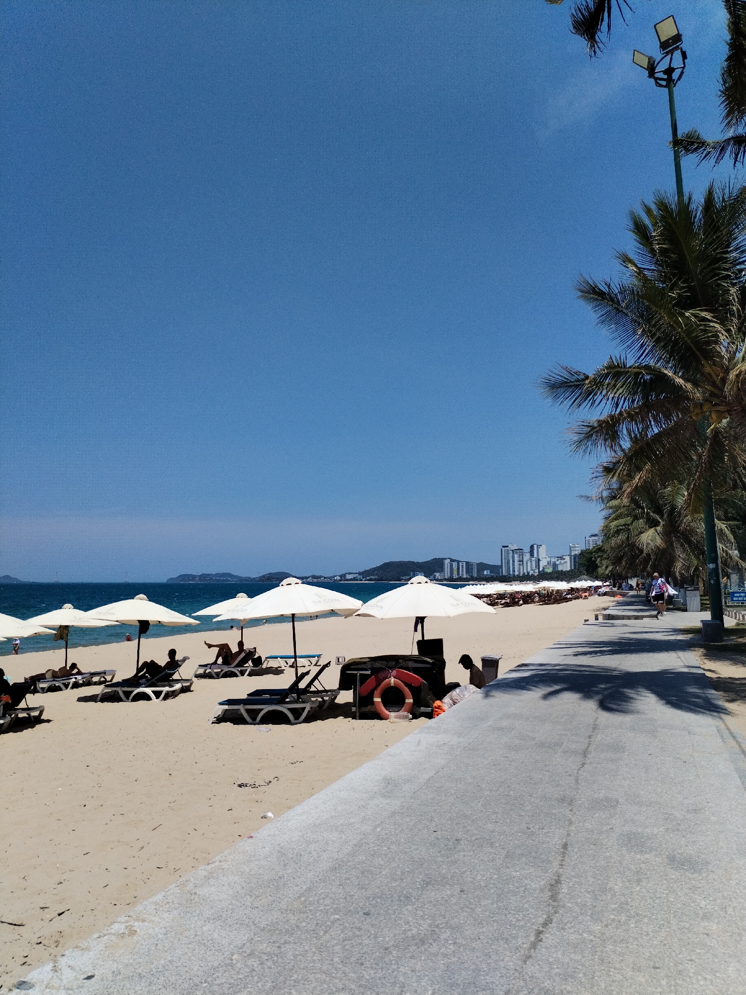 The image depicts a sunny beach scene in Nha Trang Ward, Vietnam. A row of white beach umbrellas shades beachgoers on lounge chairs on a sandy beach. Beyond the beach is a body of water with distant islands. A concrete walkway runs parallel to the beach, lined with palm trees. A woman walks along the walkway. A lifeguard ring is visible near the umbrellas. In the background, city buildings are visible. The sky is clear blue.