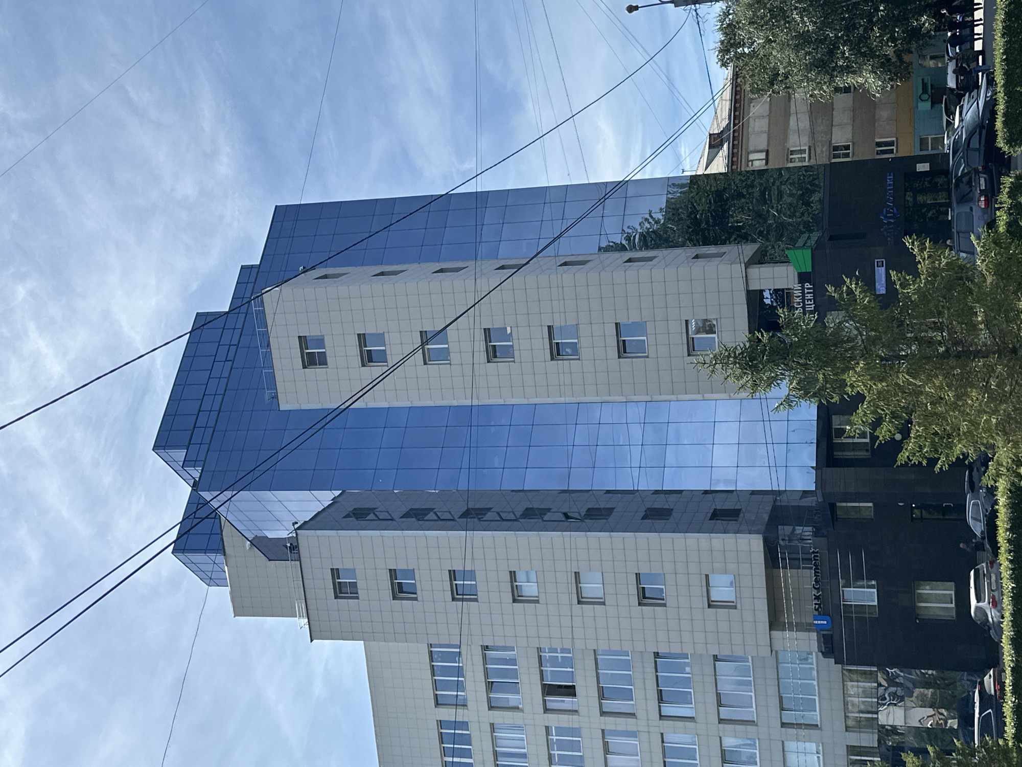 A modern, multi-story building with a facade of light-colored panels and blue glass dominates the frame. The building features a stepped architectural design, with sections of varying heights and depths. The blue glass sections reflect the sky and surrounding environment. The lower portion of the building is clad in dark, possibly black, material and has an entrance. Signs with text are visible on the lower dark facade. In front of the building, a line of cars is parked. Trees and greenery are present in the foreground and partially obscure the lower sections of the building. Overhead, utility wires crisscross the sky, creating a web-like pattern against the cloudy blue sky. The overall impression is an urban scene on a bright, possibly sunny day.