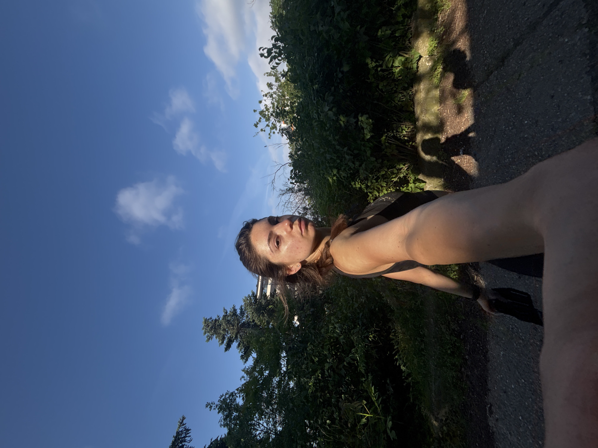 A young woman with dark hair pulled back is captured in a selfie-style photograph outdoors during daylight. She is looking towards the camera with a neutral expression. She is wearing a dark, sleeveless top. Her right arm is extended, holding what appears to be a black fabric object, possibly a mask or garment, and her hand is partially in view, obscuring the lower portion of the image.
The background consists of lush green foliage and trees, suggesting a natural or park-like setting. Behind the greenery, parts of a modern building with white and glass elements are visible, indicating an urban or suburban environment. The sky above is a clear blue with scattered white clouds. The lighting suggests it is either early morning or late afternoon, with strong shadows cast on the ground, including the woman's silhouette and that of another figure. The ground appears to be a dark, textured surface, possibly asphalt or gravel. The image is taken from a low angle, with the woman's arm and the ground occupying the foreground.
The location is the city of Varna, Bulgaria.