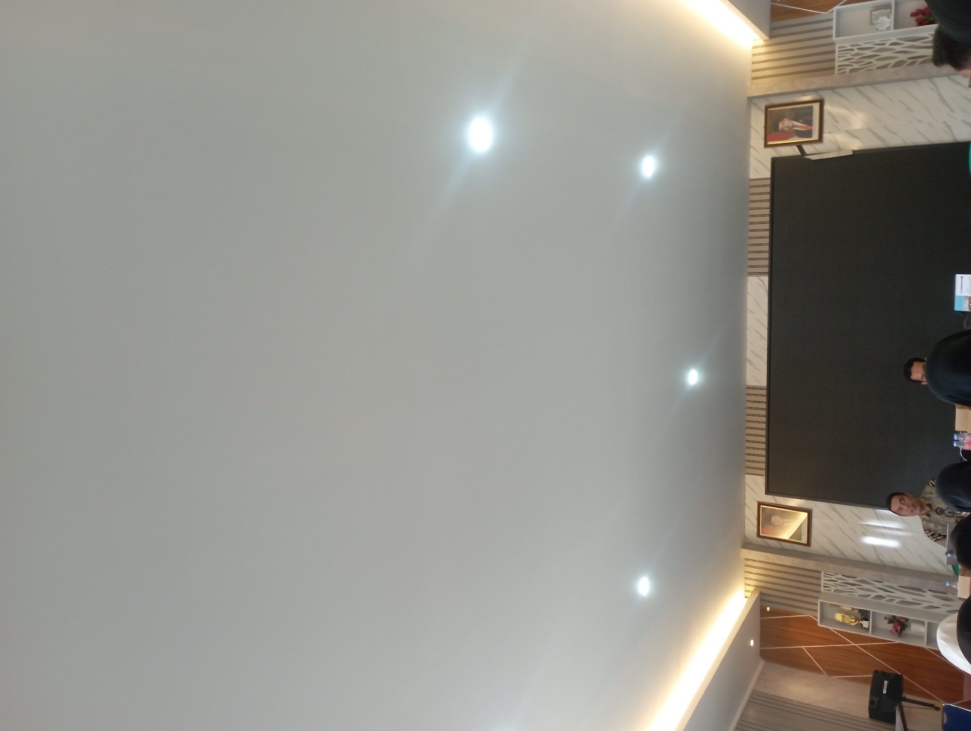 In Garut, Indonesia, a dimly lit conference room is captured from a low angle, focusing on the ceiling and the upper portion of the room. The ceiling is a plain, light gray, illuminated by several recessed lights and a warm, horizontal strip light along the top of the far wall. Two framed portraits are visible on the wall behind a large, black screen, suggesting a formal setting. One portrait on the left features a woman, likely a historical figure or politician, while the one on the right shows a man in formal attire.
In the foreground, a speaker, a man wearing a patterned batik shirt, is seated at a table, facing an audience whose heads are visible from behind. He is gesturing with his hands as he speaks, with a laptop and water bottles on the table before him. To his right, another person is partially visible, also facing forward. To the left of the speaker, a white bookshelf unit with decorative cutouts holds trophies and a small floral arrangement. A black speaker is mounted on a stand to the far left of the frame. The overall ambiance is professional and slightly muted due to the lighting conditions.