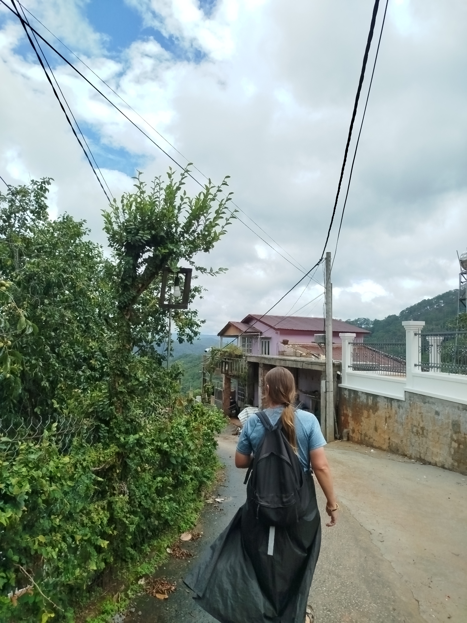 A person with long blonde hair, wearing a blue t-shirt and a black backpack, walks away from the camera down a paved road. The person is wearing a long black garment that drapes over their legs. The road is lined with a concrete wall on the right and dense green foliage on the left. In the background, there are houses with pink and beige roofs, and the scene appears to be in a hilly or mountainous area. Power lines are visible crisscrossing the sky. The sky is partly cloudy. The location is identified as Da Lat, Vietnam.