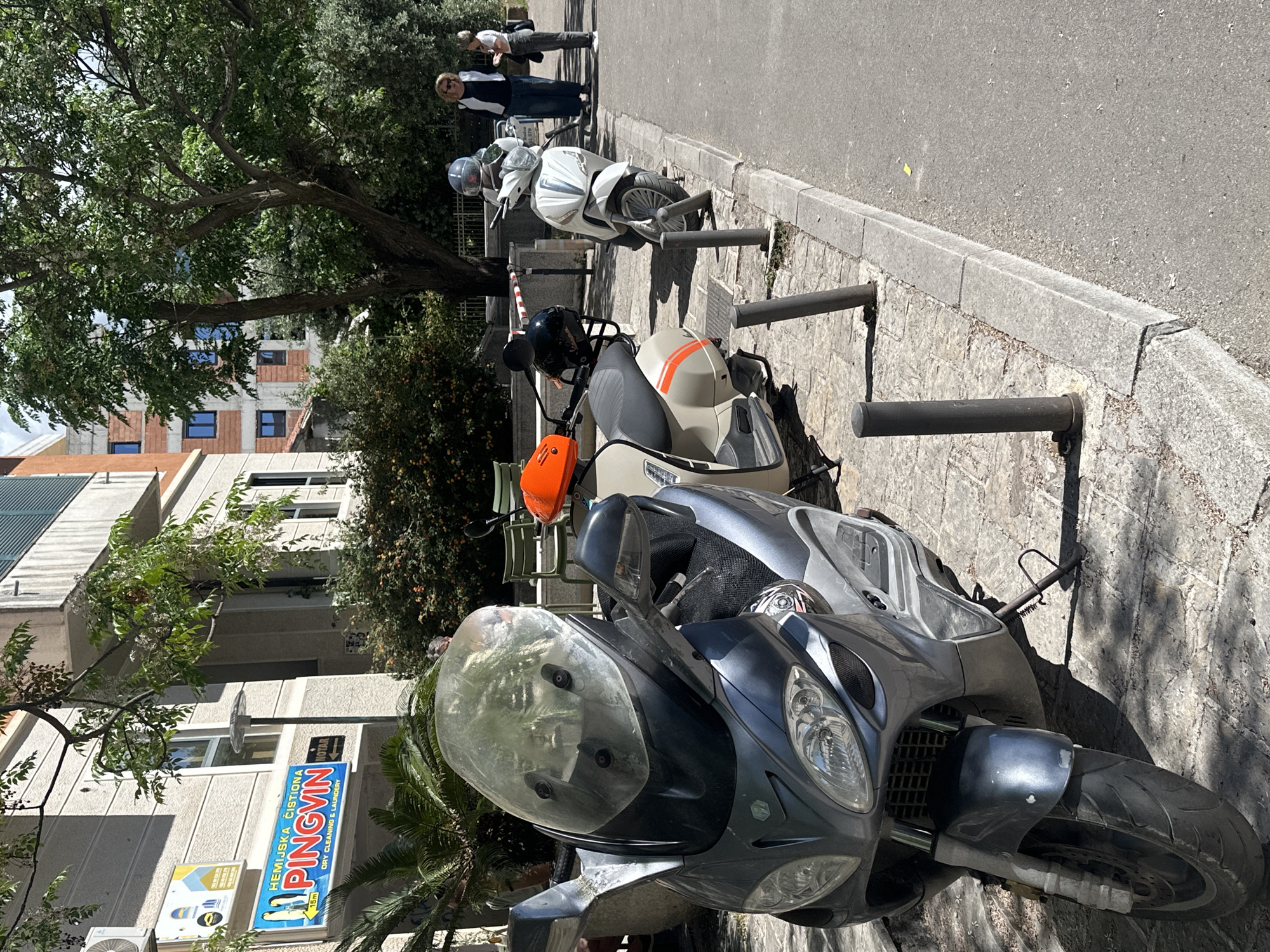 The image shows several parked scooters on a street. A sign for "PINGVIN Dry Cleaning & Laundry" is visible, along with trees and a building. People are walking on the sidewalk in the background. It is located in Budva, Montenegro.