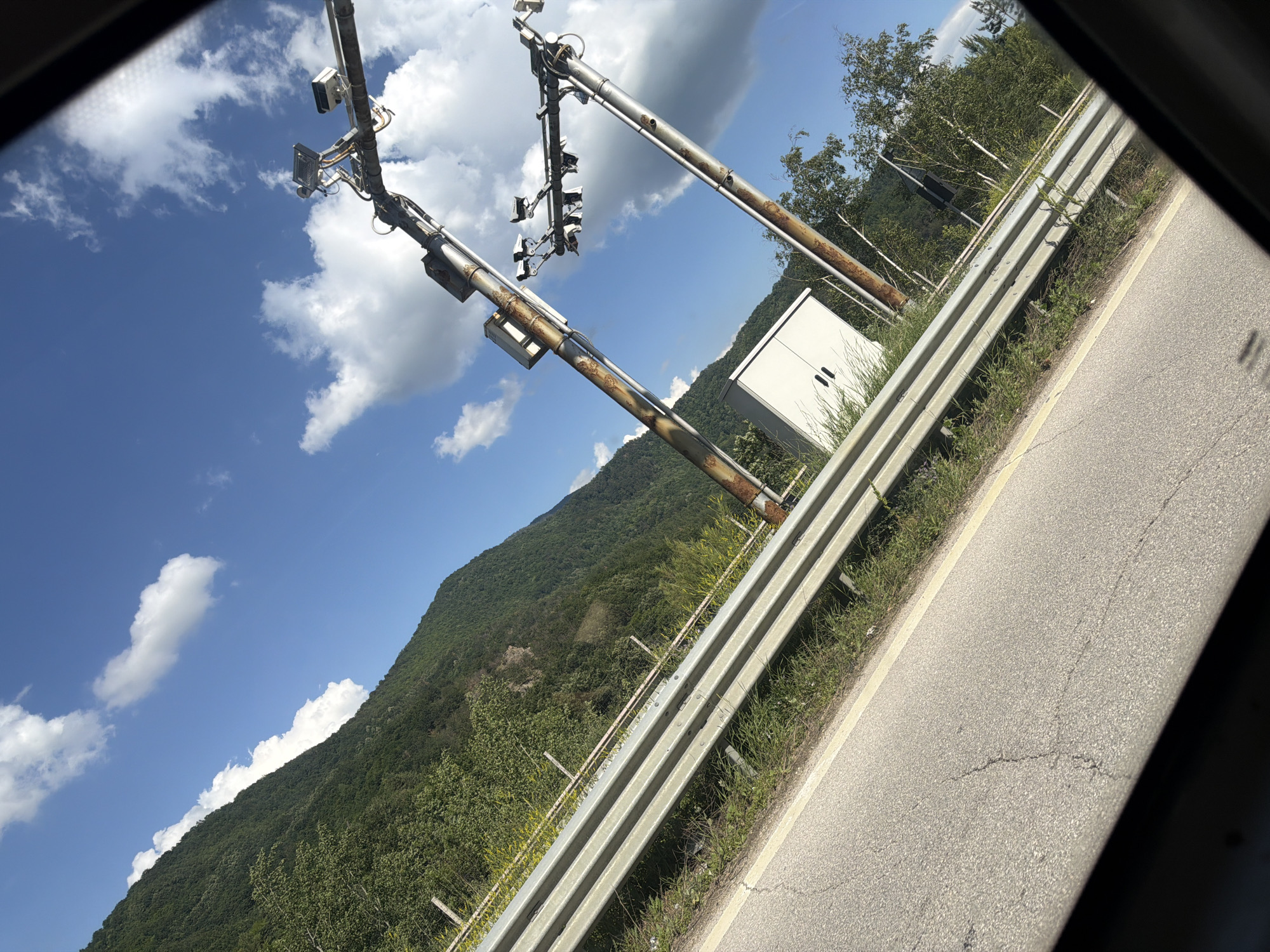 The image depicts a roadside scene in Vrachesh, Bulgaria. Two metal poles with traffic signals are prominently featured, positioned over a highway. The poles appear weathered and slightly rusted, suggesting they have been in place for some time. Attached to the poles are multiple traffic lights and what appear to be sensors or cameras, indicating an intelligent transportation system.
To the right of the poles, a white, rectangular electrical cabinet is visible, likely housing control equipment for the traffic signals. The highway itself runs diagonally across the frame, with a metal guardrail separating it from the grassy embankment. The embankment is covered in green vegetation and some wildflowers.
In the background, a densely forested mountain slope rises steeply, its greenery contrasting with the bright blue sky and scattered white clouds. The lighting suggests it is daytime, likely late morning or early afternoon, with clear and sunny weather. The perspective is from within a vehicle, looking outwards through a window, which is indicated by the dark framing on the edges of the image.
No people are visible in the image. The scene is quiet and appears to be a typical view from a vehicle traveling along a mountainous highway. There is no visible text in the image.