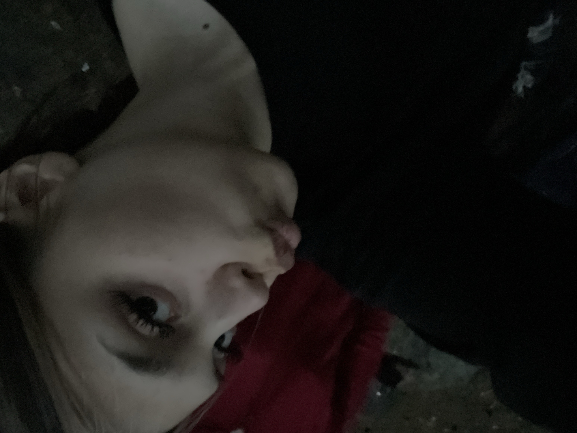 The image displays a close-up, angled portrait of a young person. The subject's face is positioned towards the viewer, featuring light-colored hair, prominent dark eyelashes, and puckered lips. A dark mole is visible on the subject's neck/shoulder area on the right. The individual wears a black long-sleeved garment, partially visible on the right side of the frame. A distinct red fabric, likely a garment or upholstery, occupies the left side of the image. The background is uniformly dark and indistinct, with scattered faint white specks suggestive of dust or very distant light reflections. There are no visual elements within the image to confirm a specific location in the city of Boitsovo, Russia.