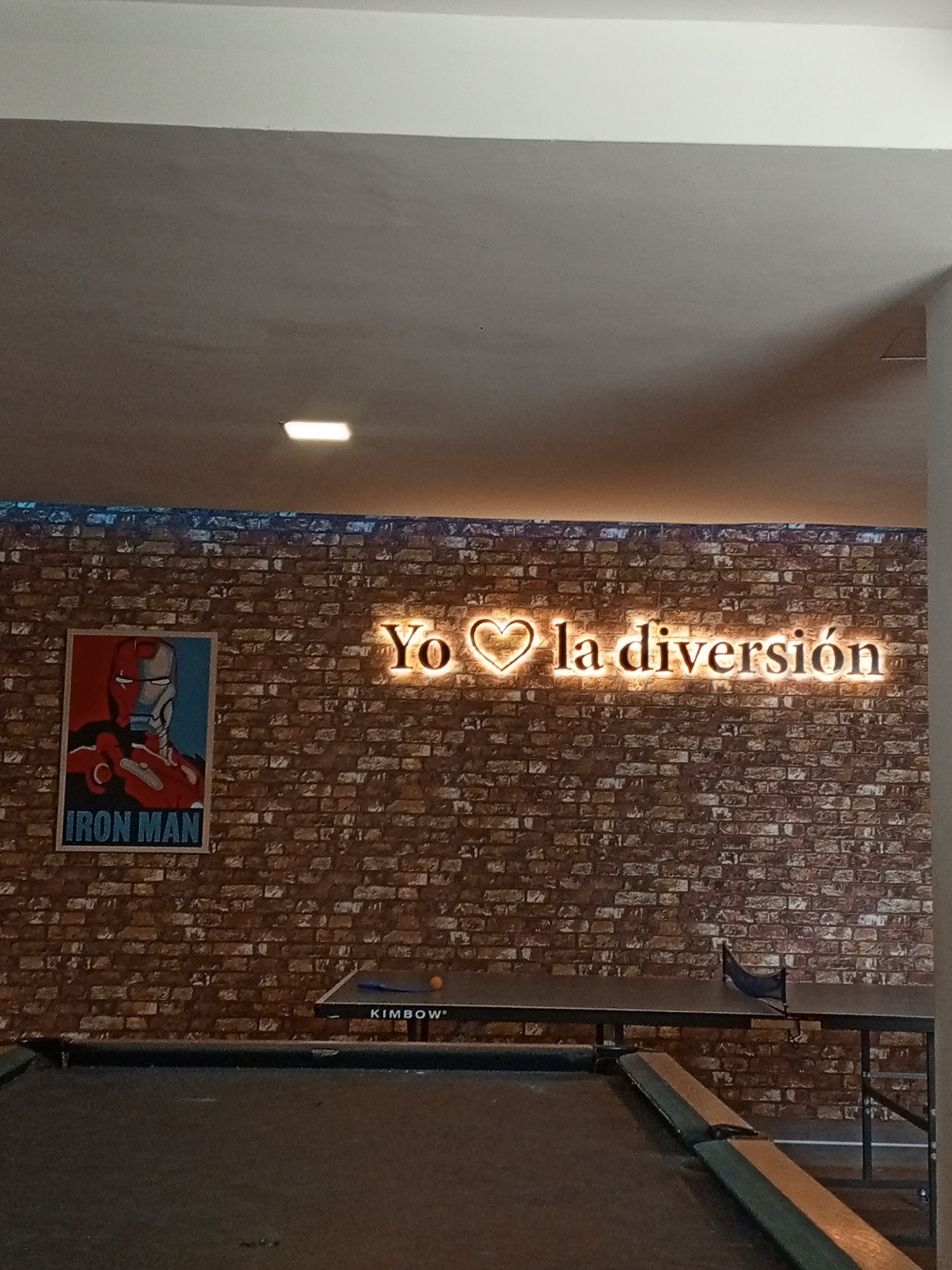 The image depicts an indoor recreation area. A pool table occupies the foreground. Behind it, a ping pong table is set up with a net and a single ping pong ball visible. A brick wall serves as the backdrop. On the wall, a poster of Iron Man is displayed to the left, and a neon sign that says "Yo ❤ la diversión" (meaning "I love the fun") in Spanish is illuminated on the right. The room is well-lit, with the ceiling appearing to be beige. Based on the Spanish phrase, the location may be in a Spanish-speaking country. The image likely represents a recreational space in Porlamar, Venezuela.