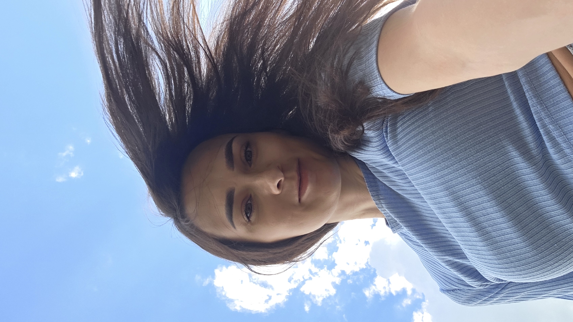 A low-angle, close-up selfie of a woman against a bright blue sky with scattered white clouds. The woman has long, dark brown hair that is being blown to her right by the wind. She is looking directly at the camera with a slight smile. She is wearing a light blue, ribbed, short-sleeved shirt. The image appears to have been taken outdoors, possibly in Дарьино, Russia, on a sunny day. The background is dominated by the sky, with no urban or landscape features visible.