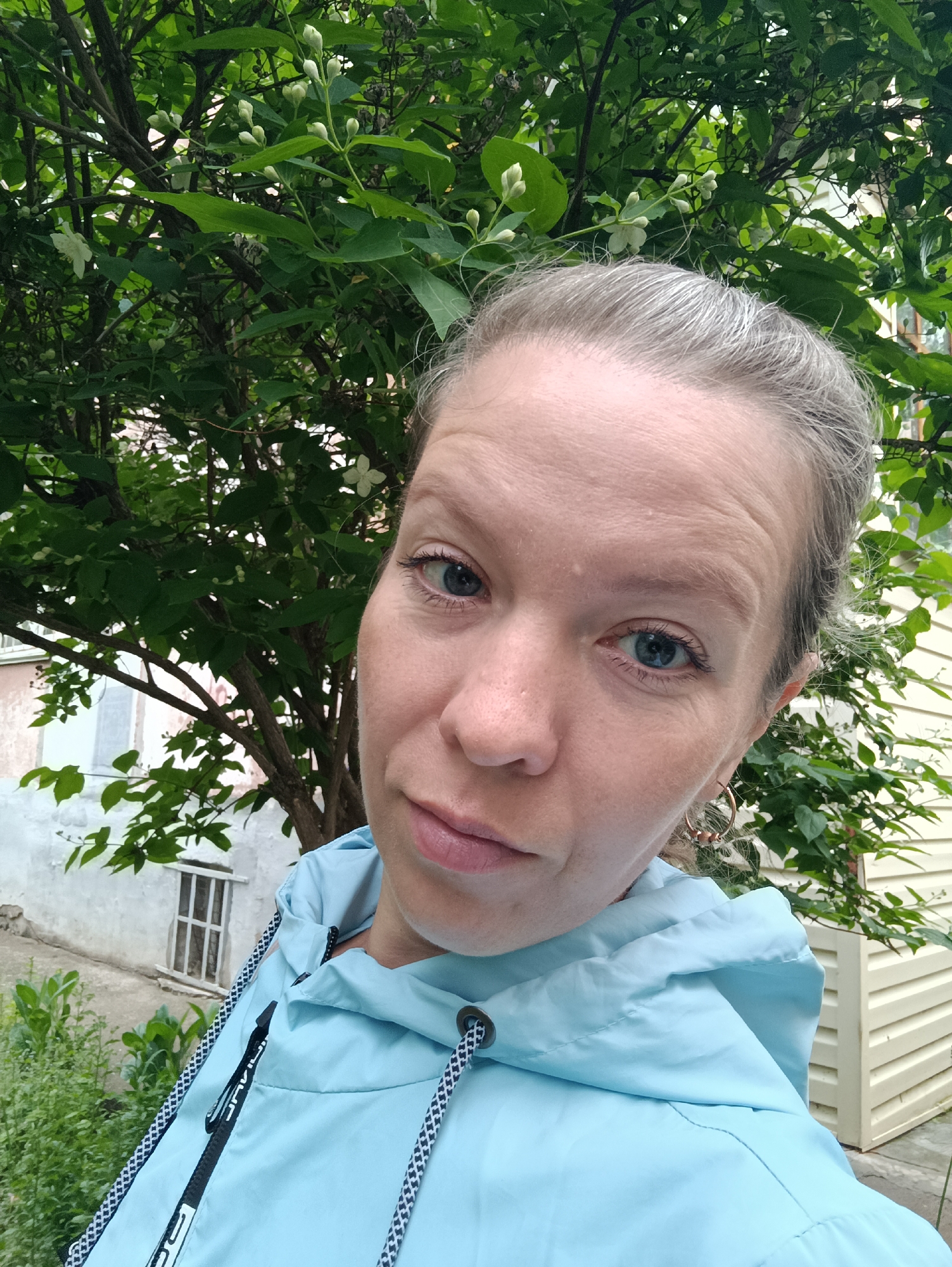 The image presents a close-up shot of a Caucasian woman with fair skin and blue eyes. She has light, possibly graying, hair pulled back. She is wearing a light blue hooded jacket with black and white patterned drawstrings. Her expression appears somewhat skeptical or questioning. She is also wearing hoop earrings.
The background features a tree with green leaves and white blossoms, suggesting it may be spring or early summer. Behind the tree, there's a building with a visible window. A light-colored, possibly wooden structure or wall is partially visible on the right.
Based on the location provided, Chaikovsky, Russia, the architecture and vegetation are consistent with what one might expect in that region. The scene appears to be outdoors in a residential or garden area. There are no specific details available to place this scene in a particular district of the city of Chaikovsky.
