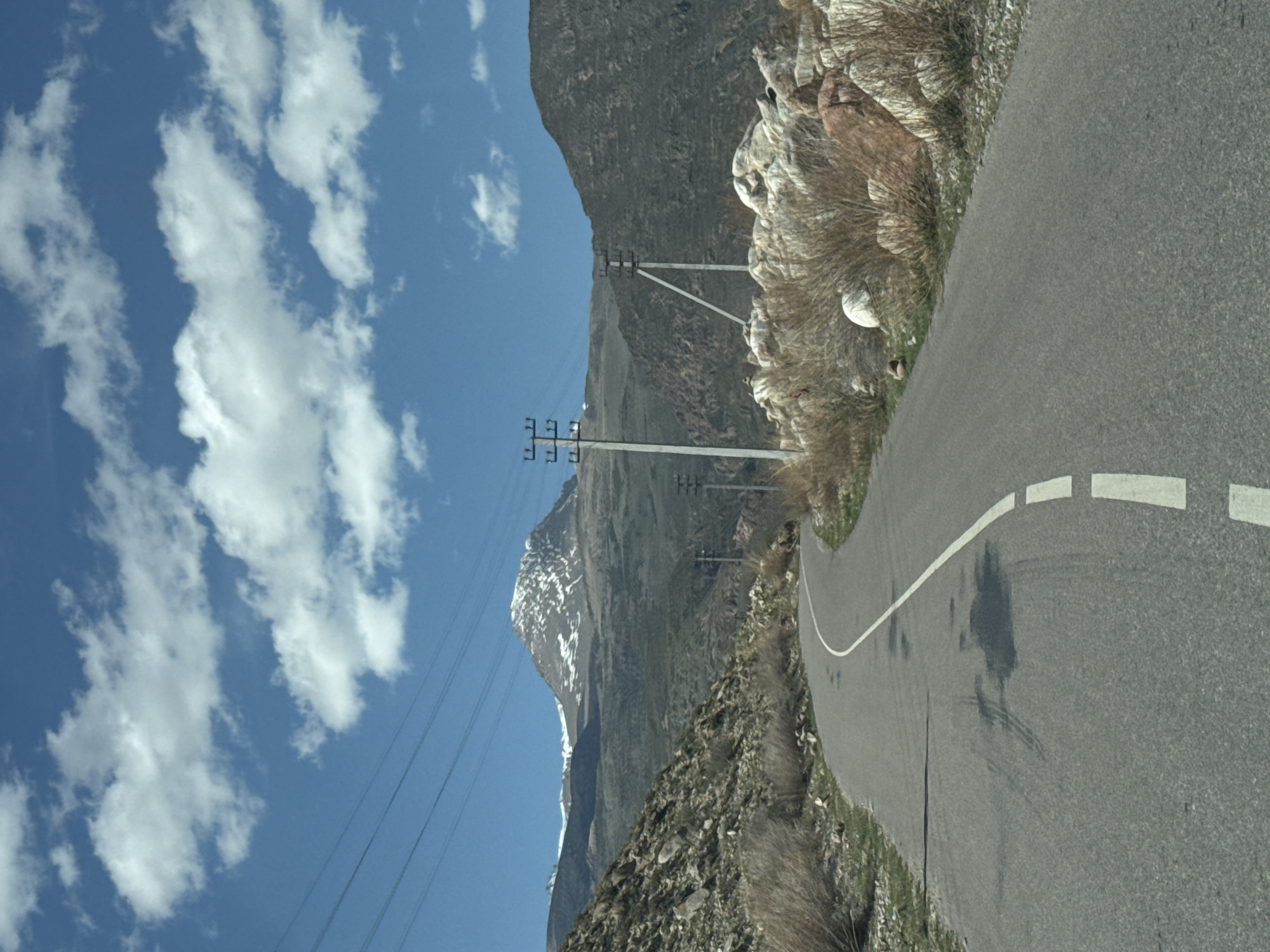A paved road with a white dashed line marking the edge curves through a mountainous landscape. The road is surrounded by rocky terrain and dry vegetation on one side, and a steep mountain slope on the other. Power lines are strung along the roadside, supported by utility poles. In the distance, a snow-capped mountain peak is visible against a clear blue sky with scattered white clouds. The image suggests a rural or remote location, possibly in a mountainous region like the outskirts of Çolpon-Ata, Kyrgyzstan. There are no people or moving vehicles visible.