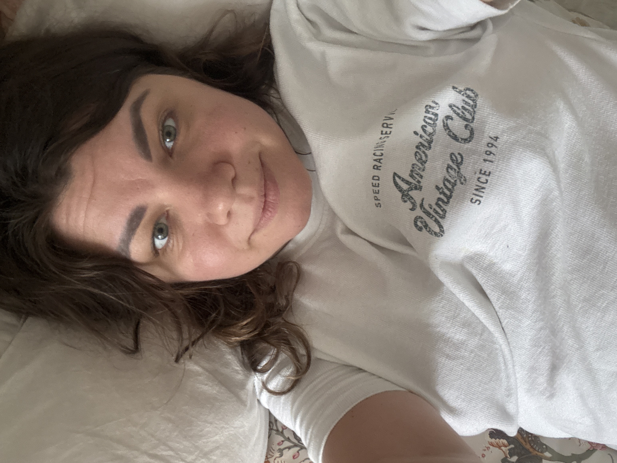 The image shows a young woman lying on a bed, taking a selfie. She has brown hair and green eyes, and she is wearing a white t-shirt with the words "American Vintage Club Since 1994" printed on it. The background appears to be a bedroom, with a pillow and a floral-patterned sheet visible. The lighting suggests it is daytime. The scene is indoors, within a residential setting. The subject is alone in the frame. The woman is positioned in the center of the image, with her face and upper body visible. Her expression is neutral. The image does not provide any information about the location being in Höhenkirchen-Siegertsbrunn, Germany.
