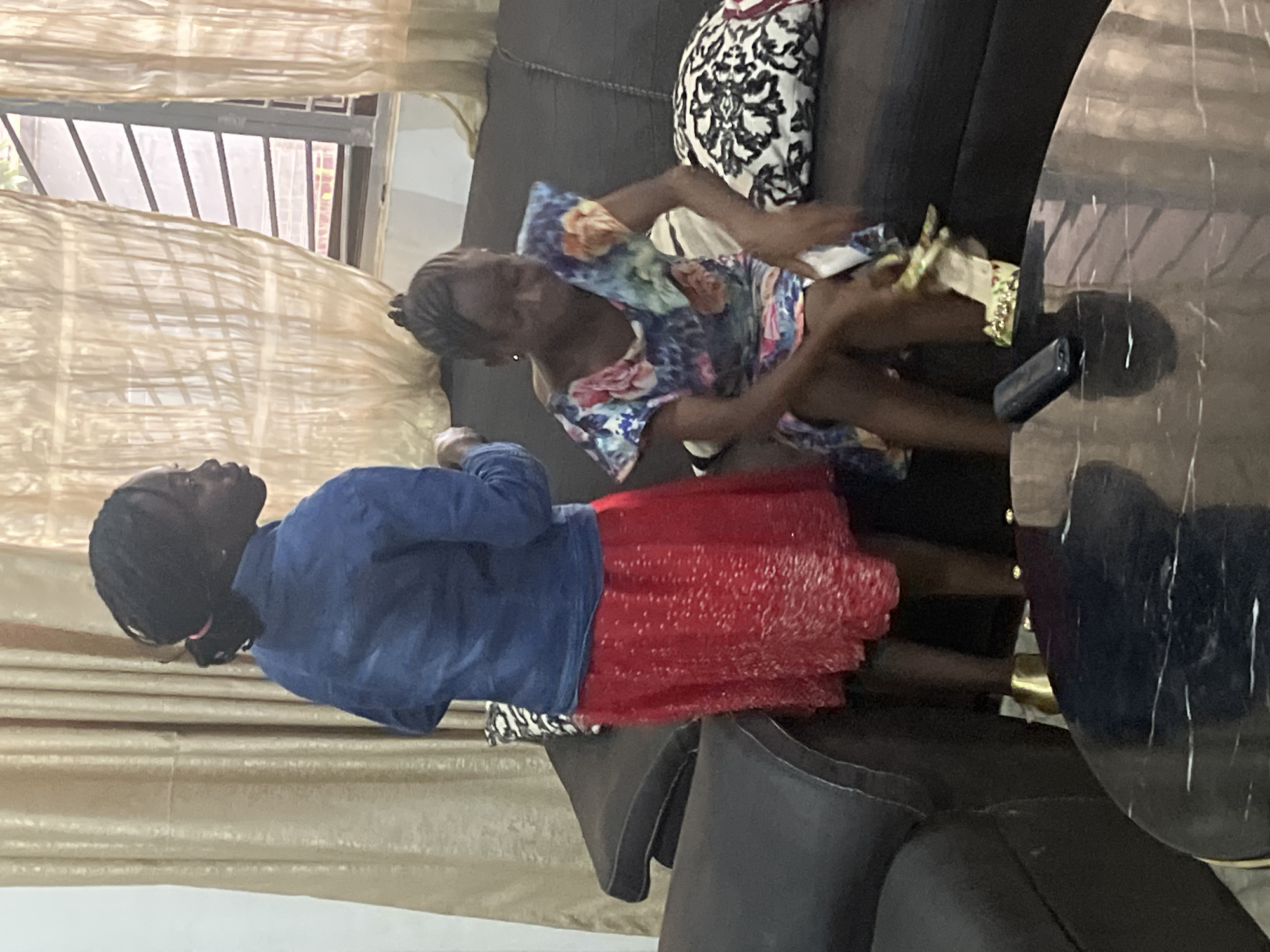 In Jos, Nigeria, two young girls, likely sisters, are seen relaxing on a couch in what appears to be a living room. The girl on the left is standing, wearing a blue denim jacket and a vibrant red skirt. Her hair is styled in braids, and she is looking upwards and to her right. The girl on the right is seated, dressed in a floral print dress. She is leaning forward, her right arm extended as if reaching for something. Her hair is also in braids. A black cell phone is resting on a marble-top table next to her. The room is decorated with sheer, golden curtains that cover a window with metal bars. A patterned cushion with black and white floral designs is also visible on the couch. The overall lighting suggests it is daytime, and the indoor setting implies a casual and familiar atmosphere.
