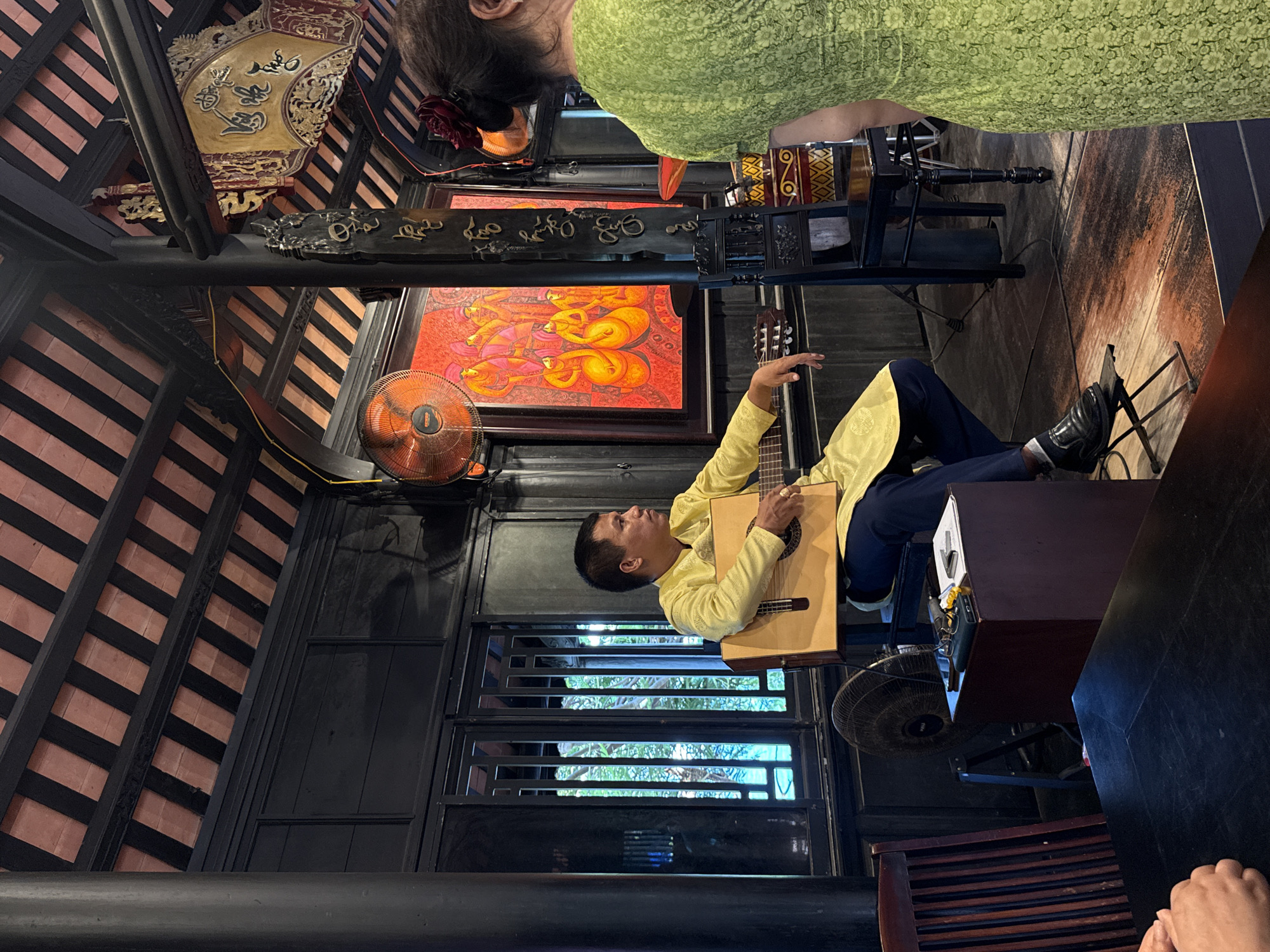 A medium shot of a musician playing a guitar in a building in Bac Nha Trang Ward, Vietnam. The musician is wearing a yellow shirt and blue pants. He is sitting on a stool and playing a guitar. There is a painting on the wall behind him, as well as two fans. The building has a wooden frame and the room is dark, with light coming in from a window. A person wearing green and with a red flower in their hair is in the top of the frame as well.
