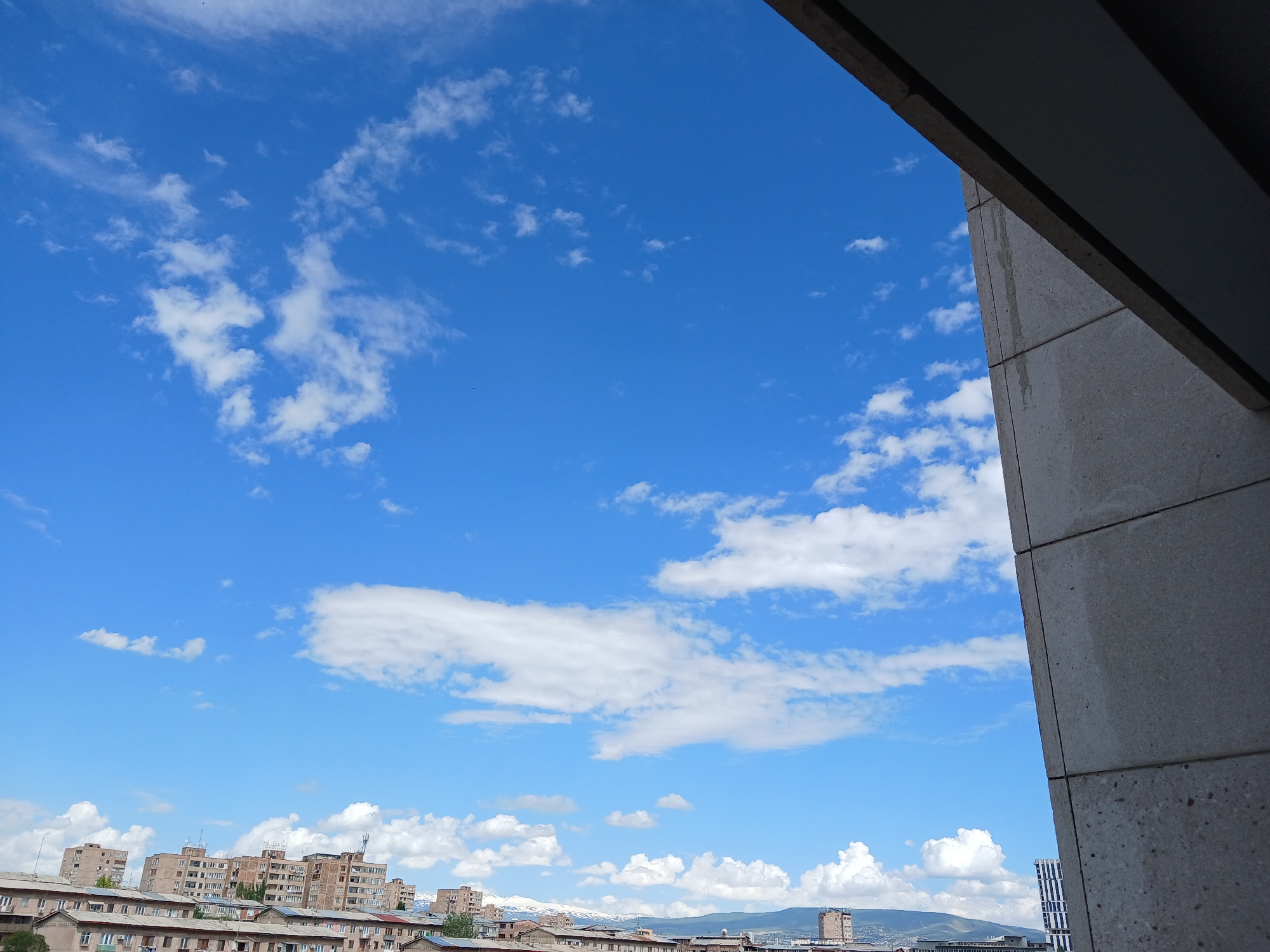 The image displays an expansive outdoor view, predominantly featuring a bright, clear blue sky adorned with scattered white clouds of varying densities and shapes, ranging from wispy cirrus-like streaks to fluffy cumulus formations.
A prominent architectural element, a large, angular, grey structure made of paneled concrete or stone, frames the scene on the right side and along the top right edge. This structure, possibly part of a building's facade or a balcony, partially obscures the sky and the distant cityscape, and shows some vertical water stains.
Below the vast sky, the lower third of the image captures a sprawling cityscape. Numerous multi-story buildings, mostly appearing to be residential apartment blocks, stretch across the horizon. Their architecture suggests a blend of styles, with some older structures visible. In the far distance, beyond the urban development, a range of mountains or hills can be seen, with distinct patches of snow visible on some peaks, particularly towards the left-center.
The time of day is bright daylight, likely mid-morning to afternoon, given the clear illumination of the scene. The weather is fair and pleasant, characterized by the blue sky and scattered clouds. No specific activities or events are taking place; it is a static landscape view. The presence of snow-capped mountains behind the city suggests a location with a temperate or continental climate, possibly captured during spring or early summer. There is no visible text in the image. The perspective suggests the photo was taken from an elevated position, looking out over the city and beyond.