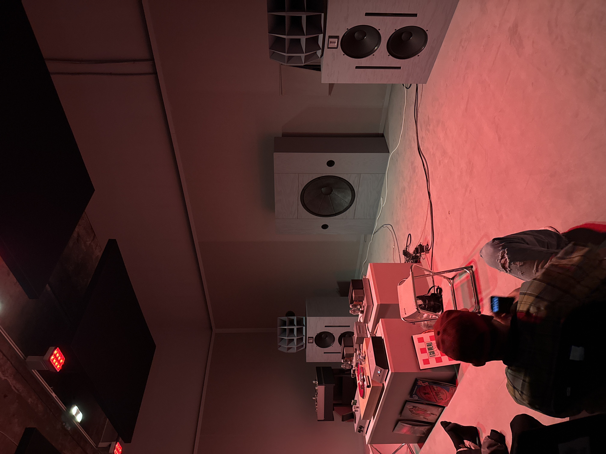A high-angle view shows an audio exhibit, bathed in reddish-pink light, likely indoors. Large, boxy speakers are mounted on the wall, some with single speakers and others with dual speakers and horn-shaped attachments. A turntable and audio equipment are arranged on a white table. A person wearing a red baseball cap and a green plaid shirt is holding a smartphone and appears to be taking a photo of the exhibit. Other people are visible in the foreground, possibly observing the setup. The overall scene suggests a focus on high-fidelity audio equipment and a live demonstration. The location, London, United Kingdom, is not discernible from the image alone.