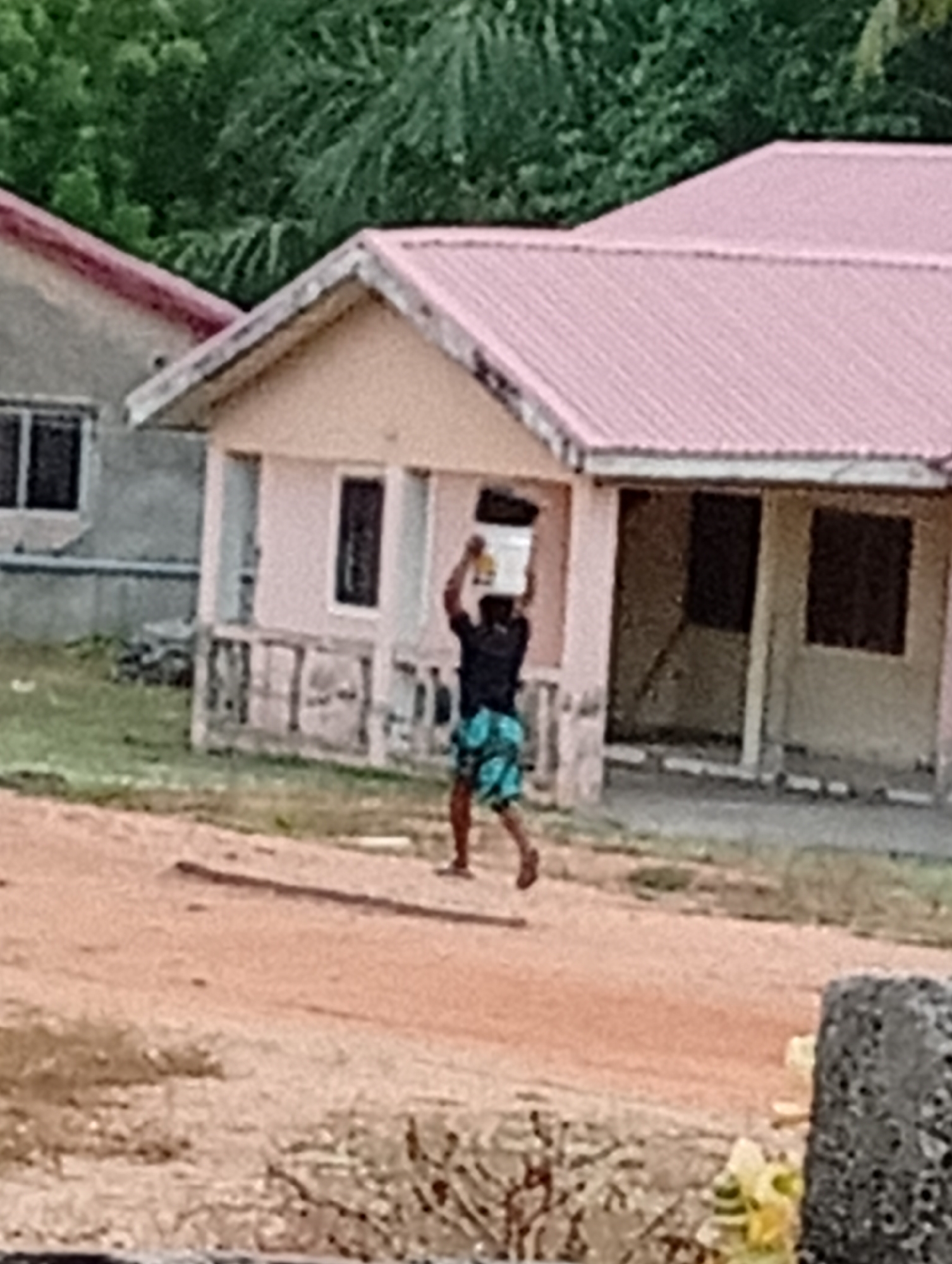 A man in a black shirt and patterned shorts is walking on a dirt road with a white bucket balanced on his head. He is walking away from the camera. Behind him is a light pink building with a metal roof of the same color. To the left of that building is another structure with a gray exterior. The background is obscured by foliage. The image appears to be taken during daylight hours. A concrete structure is in the foreground. The location is labeled Unknown, Unknown.