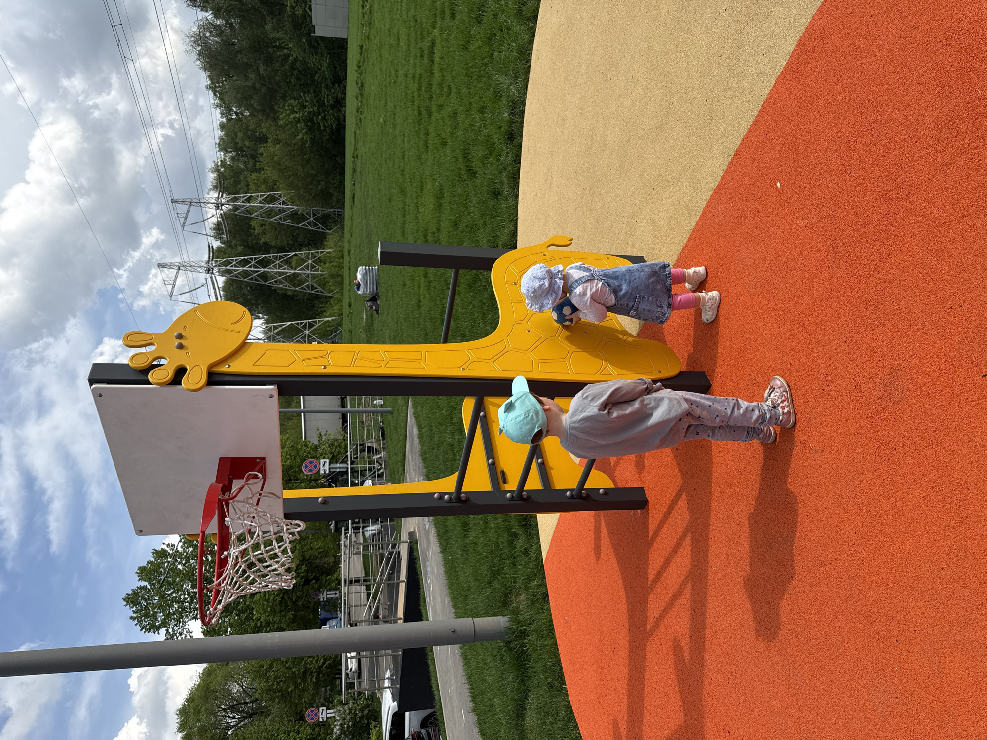 Two children play on a giraffe-shaped basketball hoop on a playground in Moscow, Russia. One child in a light blue hat leans against the black ladder, while the other, wearing a denim dress and white bonnet, holds a blue soccer ball and touches the giraffe structure. The playground has a textured, colorful surface, and a white basketball backboard with a red hoop and net is attached to the giraffe. A grassy area and trees are visible in the background, along with overhead power lines. Trees and buildings are partially visible behind the basketball hoop, along with a pedestrian walkway and parked cars.
