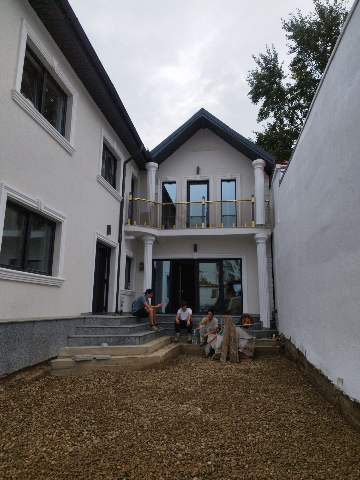 The image depicts a residential building under construction in Bucharest, Romania. The building features a modern design with white stucco walls, dark-framed windows, and decorative columns. A gravel yard is in the foreground, with construction materials visible, suggesting ongoing work. Several young men are seated on the outdoor steps, some appearing to be taking a break from their work. The sky is overcast, indicating potentially inclement weather.