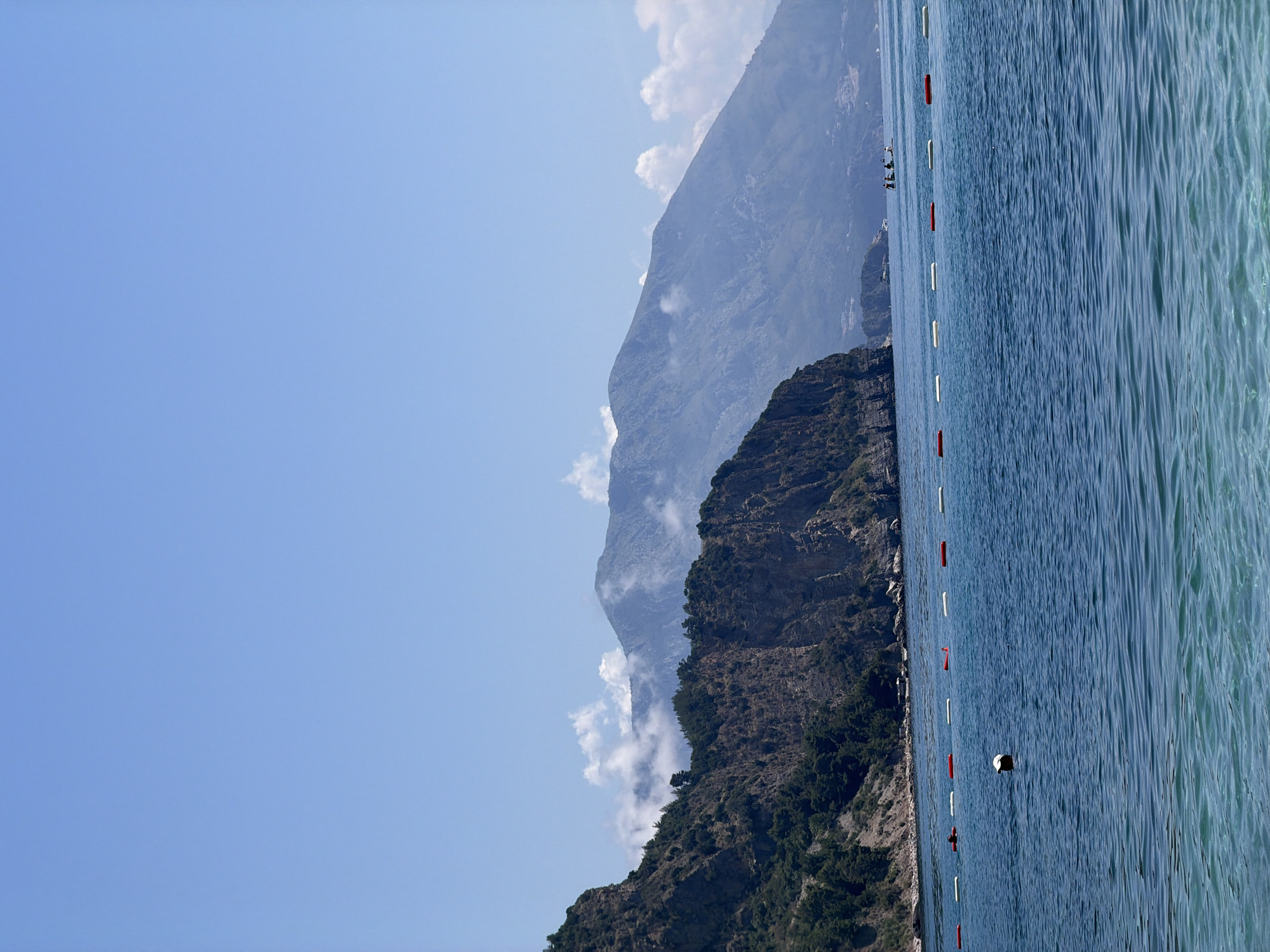 The image depicts a scenic view of the Adriatic Sea in Prijevor, Montenegro, with a backdrop of imposing mountains. In the foreground, the clear blue water shimmers, with a person wearing a hat partially submerged, appearing to swim or float. A line of buoys, alternating between red and white, demarcates a swimming area. Further out in the sea, a small group of people can be seen on what appear to be stand-up paddleboards or a small boat. The coastline is characterized by rugged, vegetated cliffs on the left, leading to larger, mist-shrouded mountains in the distance. The sky is a clear, bright blue, with a few wisps of white clouds clinging to the mountain peaks. The overall impression is one of a serene coastal landscape on a sunny day.