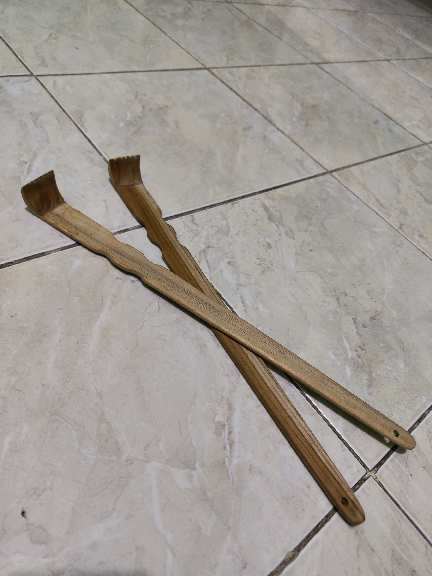 Two wooden back scratchers are placed diagonally on a tiled floor. The tiles are light-colored with a marble-like pattern and are separated by dark grout lines. The back scratchers are of similar design, each with a long, slender handle that has a slightly curved or wavy contour. The ends of the scratchers are notched, designed to provide a scratching surface. The scratchers are positioned so that their tips are towards the top left of the frame, and their handles extend towards the bottom right. The lighting suggests an indoor environment, possibly a home or shop. The location is specified as Sroyo, Indonesia.