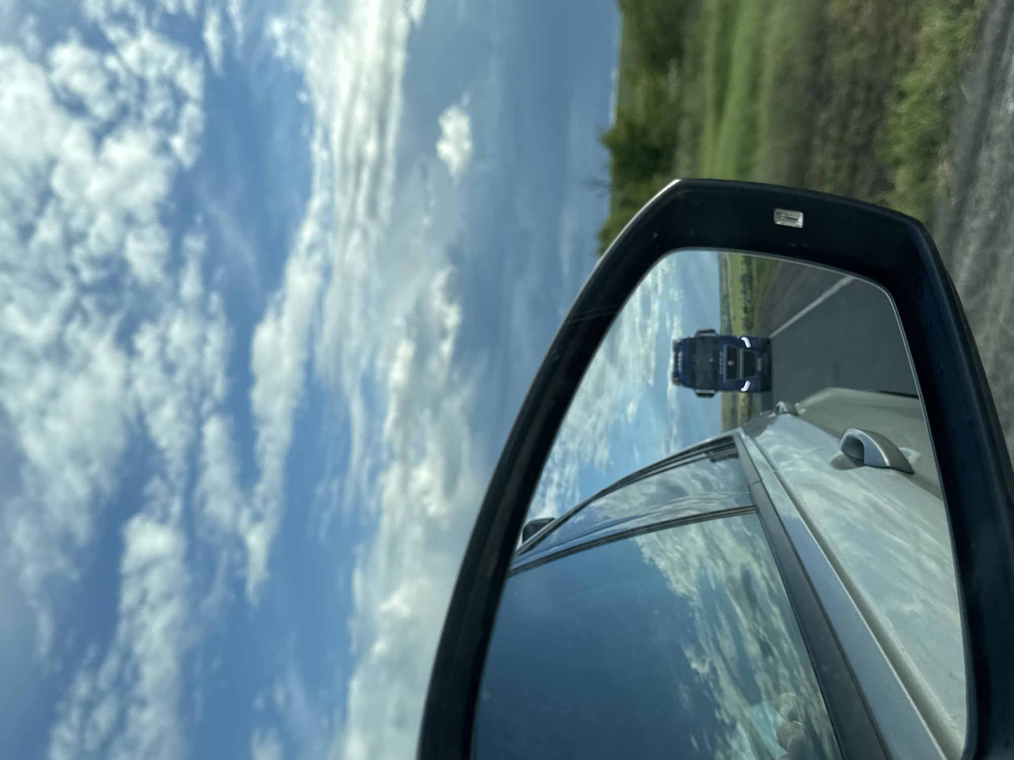 The image captures a view from inside a moving vehicle, likely in Belogorki, Russia, with a significant portion dedicated to the sky and the car's side mirror. The camera is oriented vertically, causing the natural horizon to appear on the right side of the main frame.
The central subject is the black-framed side mirror, which clearly reflects a large, dark blue or black truck following directly behind the observer's car on a paved road. The road appears straight and extends into the distance within the mirror's reflection, suggesting open terrain.
Outside the car, a bright blue sky dominates the left and upper parts of the frame, filled with scattered white cumulus clouds. This indicates a clear and sunny daytime. To the right of the mirror, green vegetation, possibly fields or unkempt grass and bushes, lines the road, suggesting a rural or semi-rural setting.
Parts of the car from which the photo is taken are visible. The silver-colored bodywork, including a side window and a roof-mounted antenna or similar fixture, reflects the sky and clouds, adding depth to the scene. The bright lighting suggests it is mid-day. There are no people visible, and no discernible text can be seen anywhere in the image. The overall impression is one of travel or a road trip under fair weather conditions.