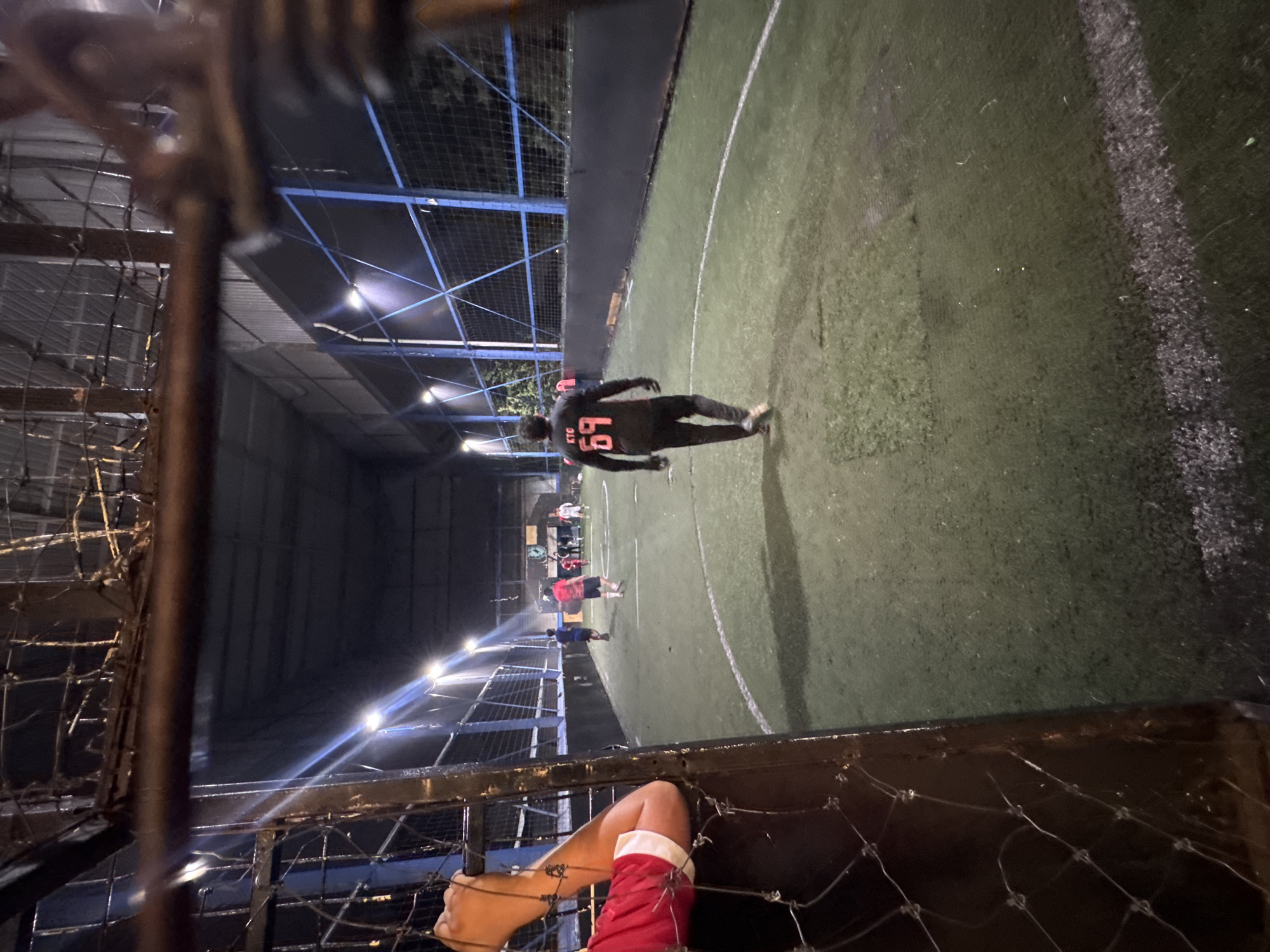 The image shows an indoor soccer field with a person wearing a black jersey with the number 69 in orange on the back. Other people are visible on the field as well. The view is from behind a chain-link fence, with a hand and part of a red and white sock visible in the foreground. The lighting is artificial, and the building has a metal frame and roof. Based on similar images found online, the location could be Puebla, Mexico.