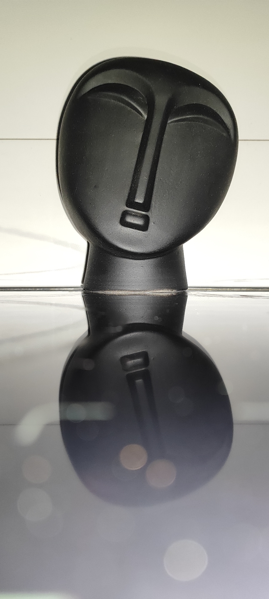 A black abstract sculpture of a face is positioned on a reflective surface, creating a mirrored image below it. The sculpture is dark, with carved features representing eyes, a nose, and a mouth. The background is a plain white surface, possibly a wall or shelf. The reflective surface below is dark and smooth, showing blurred circles of light, suggesting ambient lighting. The image is captured from a slightly elevated angle, looking down at the sculpture and its reflection. The location in Snezhinsk, Russia is not discernible from the visual information provided in the image.